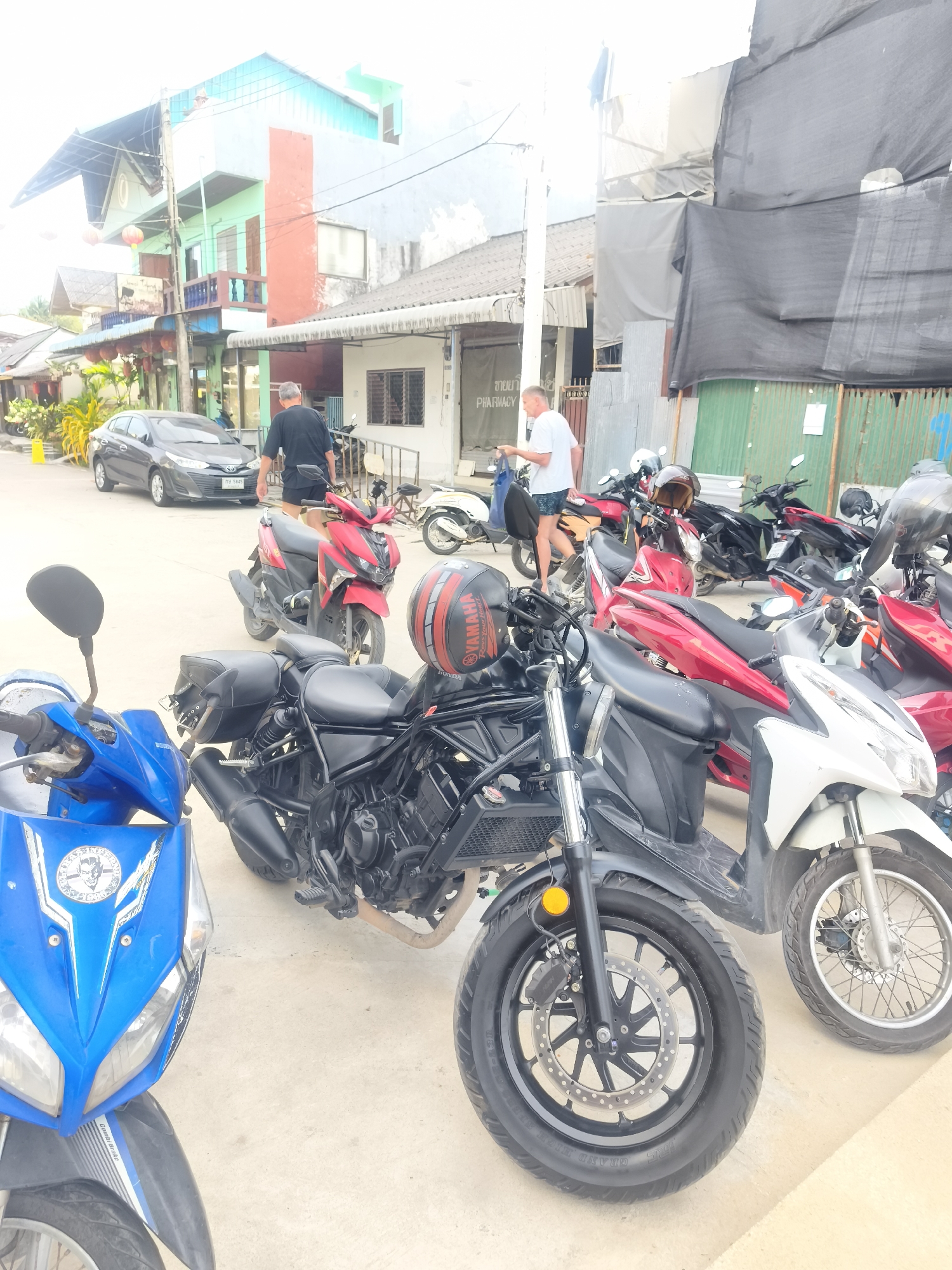 This daytime image captures a vibrant street scene in Baan Mae Nam, Thailand, filled with people, vehicles, and buildings. The lighting is bright, suggesting either full sun or a lightly overcast sky.
In the foreground and midground, a variety of motorcycles and scooters are parked. Most prominent is a black motorcycle in the center, equipped with black saddlebags, and a red and black helmet with "YAMAHA RACING FACTORY" text resting on its fuel tank. To the left, the front portion of a bright blue scooter, featuring a circular graphic sticker, is clearly visible. Numerous other scooters, primarily red and white, are parked to the right, forming a dense line of vehicles.
Several individuals are present. A man in a black t-shirt and dark shorts is walking away from the camera, heading towards a silver Toyota Yaris sedan parked on the left side of the street. The car displays a Thai license plate, "กย 9365". Another man in a white t-shirt and patterned shorts is seen on the right, carrying a blue bag, standing near the parked scooters.
The background reveals a mix of buildings. On the left, a multi-story building features a distinctive light blue and green facade with orange accents, balconies, and hanging red lanterns. Further back, more modest structures are visible, including one with a sign indicating a "PHARMACY" (with Thai script "ขายยา" also visible). Parts of the buildings on the right are covered with large grey and black tarps. The street itself appears to be paved, and a utility pole with overhead wires is visible on the left. The scene depicts a lively, everyday street environment characteristic of a Thai town or village.