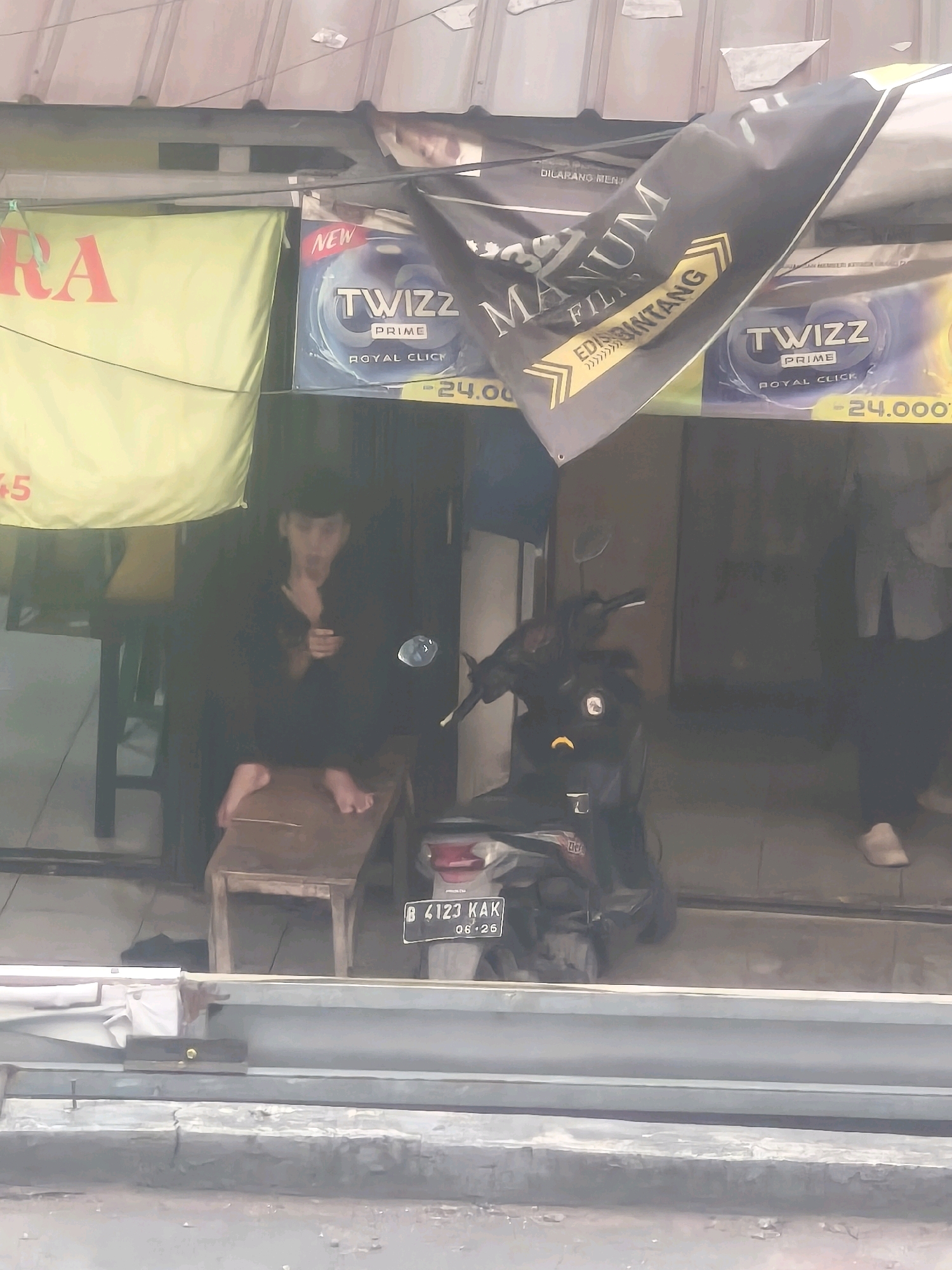 The image depicts an outdoor scene in Bekasi, Indonesia. A young man in dark clothing is seated on a wooden stool, with his feet resting on a smaller wooden platform. A black motorcycle is parked to his right. Above and behind the man, banners are displayed. One banner, partially obscured, shows "RA" in large red letters on a yellow background. Another banner, black with white and yellow text, features "MANUM FIL" and "EDIS BINTANG". To the right of this, a blue and white banner advertises "TWIZZ PRIME ROYAL CLICK" with the price "24.000". The ground in front of the motorcycle is paved.