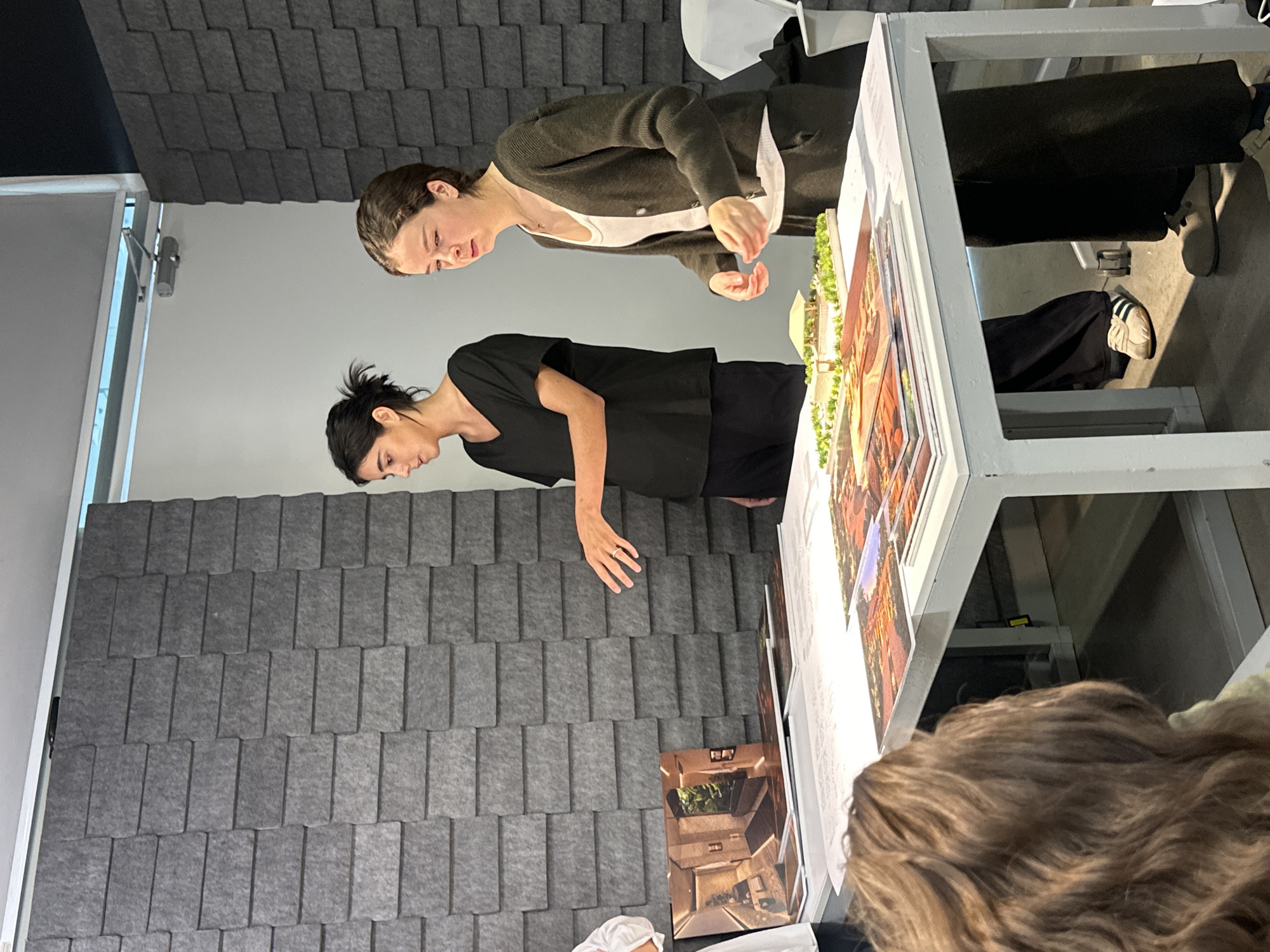 Two people stand near a white metal table with images and a model on it. One wears a black top and the other wears a white top with a cardigan. A third person's brown hair is visible in the foreground. A wall with textured gray panels is in the background. The location is likely an indoor space, possibly in Mexico City, Mexico.