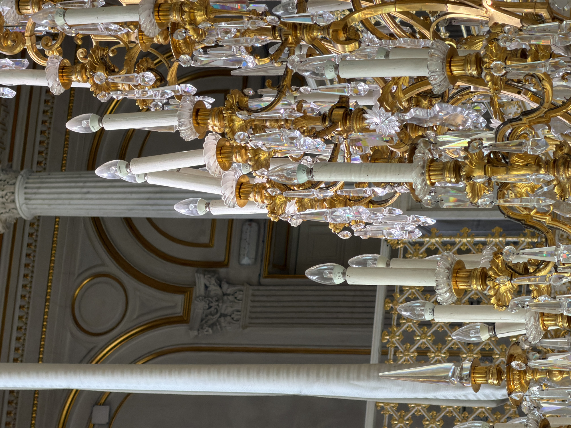 The image shows a close-up of an ornate, gold-finished chandelier with numerous glass crystal accents and white candle-shaped light fixtures. The chandelier is decorated with intricate scrollwork and floral motifs. The light bulbs are clear, conical, and appear to be LED.
The setting appears to be an interior of a grand building, possibly a palace or a historic theater in Saint Petersburg, Russia, given the ornate gold trim on the ceiling in the background. The ceiling itself is white with gold decorative moldings, including circular and rectangular patterns. A large white cylindrical column is partially visible on the left side of the image. The overall impression is one of opulence and grandeur.
No people are visible in the image. There is no indication of the time of day or weather conditions. The activity is simply the display of the chandelier in its setting.
Visible text on the light bulbs reads "CE" and what appears to be "PHILIPS".