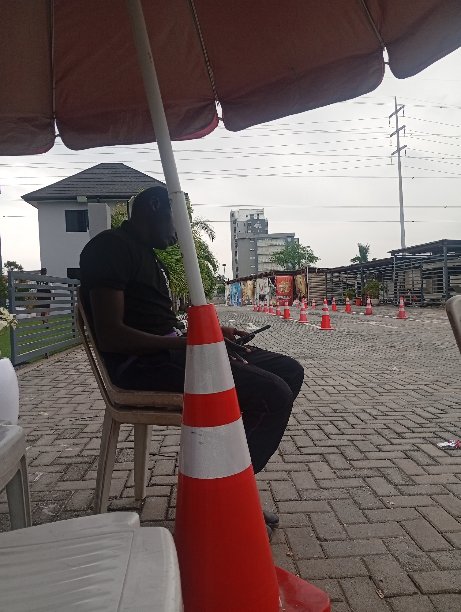 An individual, a Black male wearing a black t-shirt and dark pants, sits in a plastic chair under a large red umbrella. He is looking down at a mobile phone in his hands, his legs crossed. The chair is positioned on a paved brick surface. In the foreground, to the right of the individual, is a bright orange traffic cone with two reflective white bands. Beyond the individual, the environment consists of a paved area lined with traffic cones, leading towards a series of buildings and structures. In the background, there is a modern building with dark windows and a distinctive roofline, a tall electrical pylon with power lines, and a longer, low-rise building with an industrial appearance, possibly a warehouse or market stall structure. The sky is overcast and light. The overall setting appears to be an outdoor public space, possibly a commercial or event area, in Ilado, Nigeria.