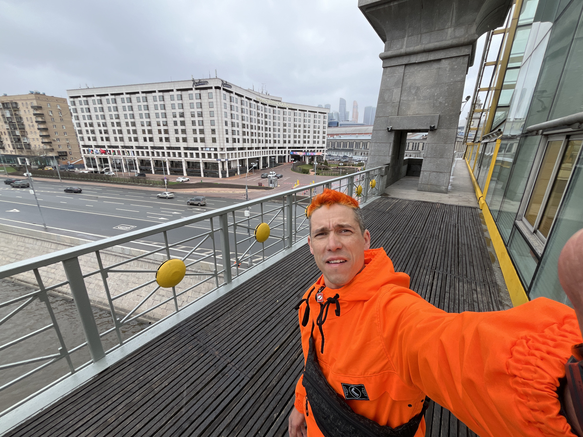 A man with orange hair stands on a bridge in Moscow, Russia, taking a selfie. He wears an orange jacket and a black shoulder bag. The bridge has a gray metal railing decorated with yellow circular accents, and a wooden plank walkway. A multi-lane road with cars runs below. In the background is a large white building, identified as the Radisson Slavyanskaya Hotel and Business Centre, and the skyscrapers of Moscow City are visible in the distance. Part of the bridge structure includes gray stone and a glass-enclosed walkway. The sky is overcast.