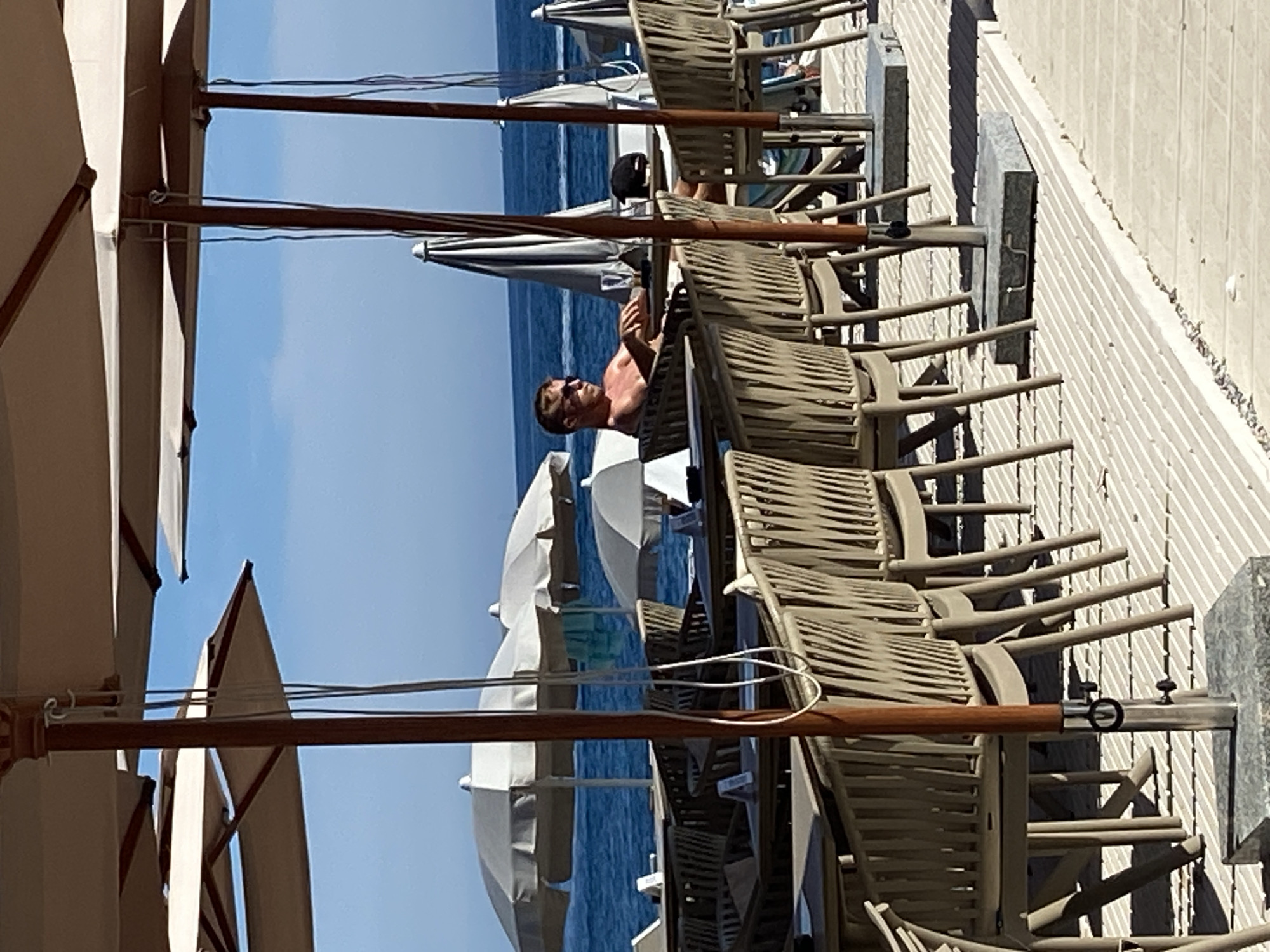 The image depicts a sunlit outdoor seating area at a beachside establishment in Budva, Montenegro. In the mid-ground, a shirtless man with blonde hair and sunglasses is seated at a table, looking towards the left. He appears to be relaxed. The seating area is furnished with numerous tables and chairs, many of which are empty. The chairs are made of woven material in a neutral tone, and the tables have light-colored tops. Large, beige umbrella structures are erected to provide shade, with their wooden poles visible. In the background, the blue ocean stretches out under a clear sky, with several white umbrellas dotted along the shoreline and a distant boat on the water. The ground is a light-colored wooden deck with a repeating linear pattern. The overall scene conveys a leisurely atmosphere characteristic of a seaside resort.