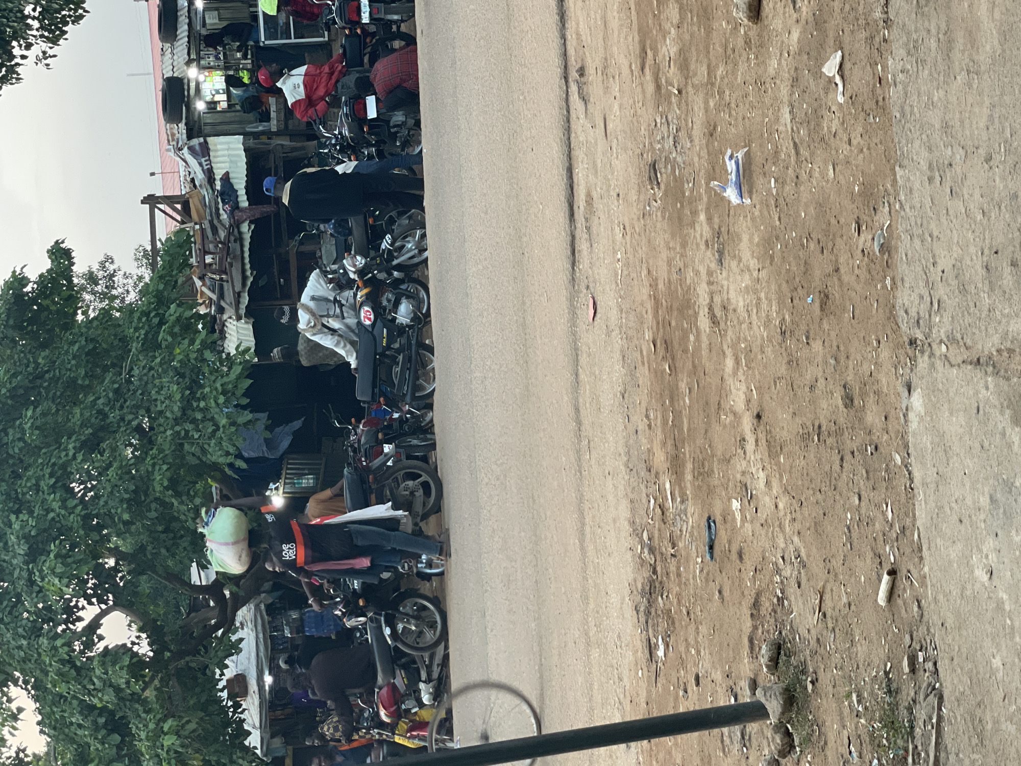 The image depicts a street scene in Barden Yana, Nigeria. In the foreground, a dusty, unpaved area borders a paved road. A dark pole stands on the left side of the frame, and debris, including rocks, plastic wrappers, and a discarded cylindrical object, is scattered across the ground.
The road itself is the focal point of activity. Parked motorcycles are lined up along the roadside, some with riders or passengers. Several individuals are visible interacting with the motorcycles or standing nearby. One person in the center is wearing a white garment and a blue head covering, standing beside a motorcycle. Another person, carrying a large, light-colored bundle on their head, is wearing a black t-shirt with orange accents and a red scarf. Their shirt has text that includes "LOVE VELO".
Behind the motorcycles, a row of market stalls or shops is visible, constructed with corrugated metal roofs and wooden supports. The stalls appear to be selling various items, including clothing and electronic goods, indicated by displays of merchandise. People are browsing or working within these stalls. A large tree with lush green foliage provides shade over the market area and part of the road. The overall atmosphere suggests a bustling, informal marketplace. The sky is overcast, indicating a potentially hazy or dull day.