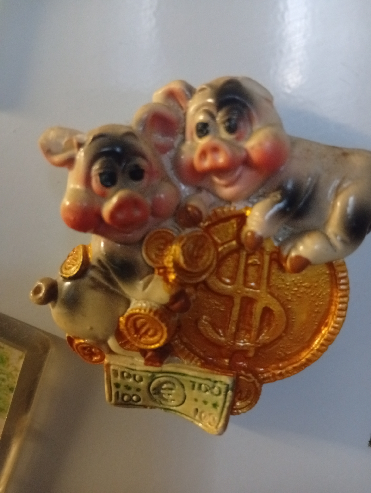 A small, decorative figurine of two smiling pigs is depicted. The pigs are light-colored with black markings around their eyes and on their backs. They are positioned atop a large, gold-colored coin with a dollar sign in the center. Surrounding the pigs are several smaller gold coins. Below the coin, there is a green-colored bill with the number "100" printed twice and a euro symbol in the center. The figurine appears to be made of a plastic or ceramic material. The background is a plain, light-colored surface, possibly a wall or refrigerator. There is no specific information about the location in the city of Pushkino, Russia, as the image does not contain any identifiable landmarks or context clues related to a specific geographical area.