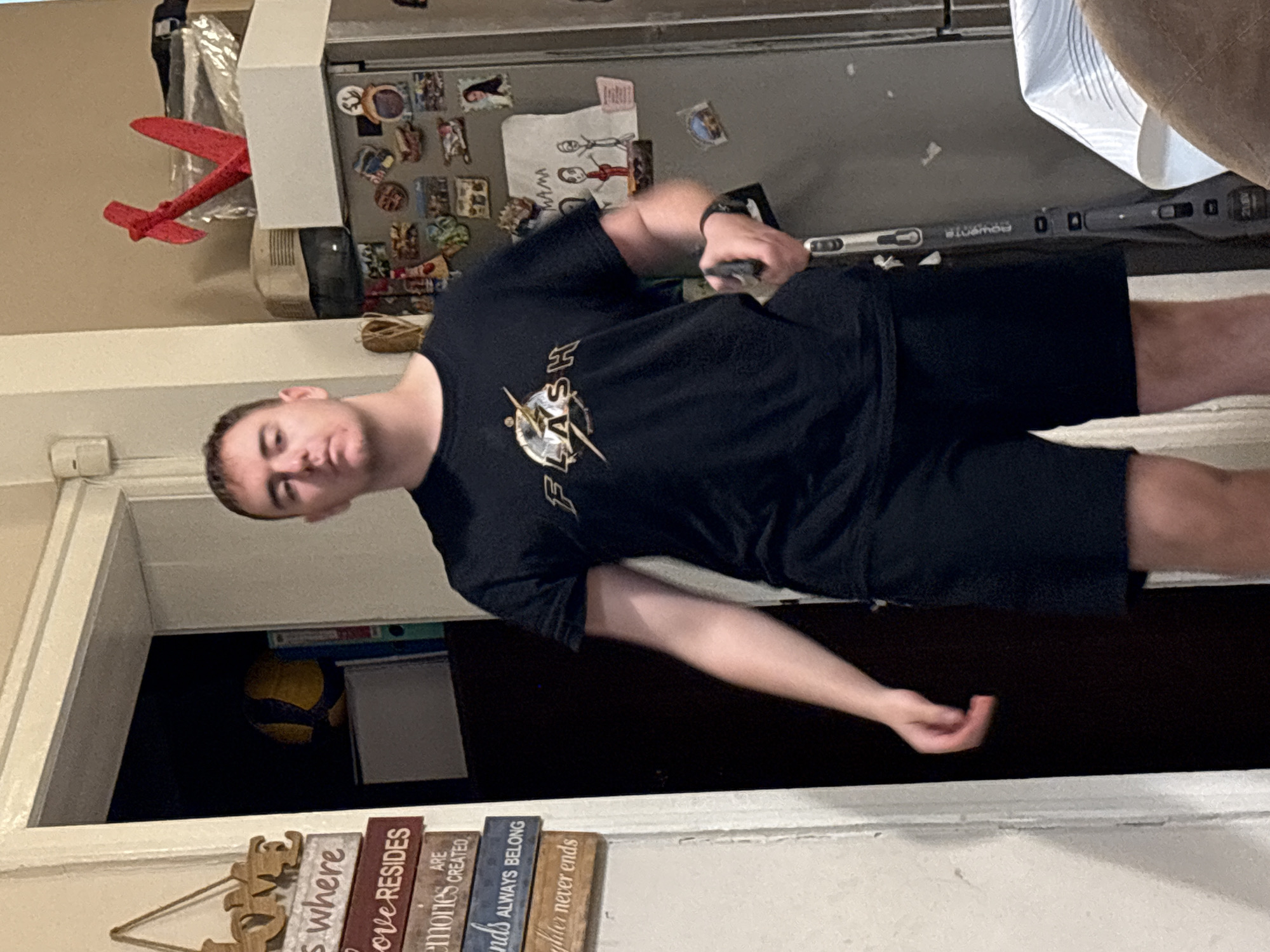A male individual is observed standing indoors in what appears to be a residential setting in Sofia, Bulgaria. The subject, an adult male with short dark hair, is wearing a black t-shirt with a gold and white "FLASH" logo and black shorts. He holds a black and silver handheld vacuum cleaner, partially obscured, in his right hand. To the left of the subject, a white wall displays stacked wooden decorative signs with phrases such as "HOME is where LOVE RESIDES," "MEMORIES ARE CREATED," "FRIENDS ALWAYS BELONG," and "laughter never ends." An open doorway or archway to the left reveals a darker room containing a yellow and blue volleyball and multiple binders (one white, one blue). To the right of the subject, a stainless steel refrigerator is adorned with numerous magnets, including photographs and a drawing labeled "MAMA" featuring stick figures. On top of the refrigerator, a white rectangular box, a red angular decorative object, and an older beige electronic device are visible. A white tablecloth partially covers a surface in the bottom right, and a portion of brown upholstered furniture is also visible. The environment appears to be an interior domestic space, possibly a kitchen or living area.