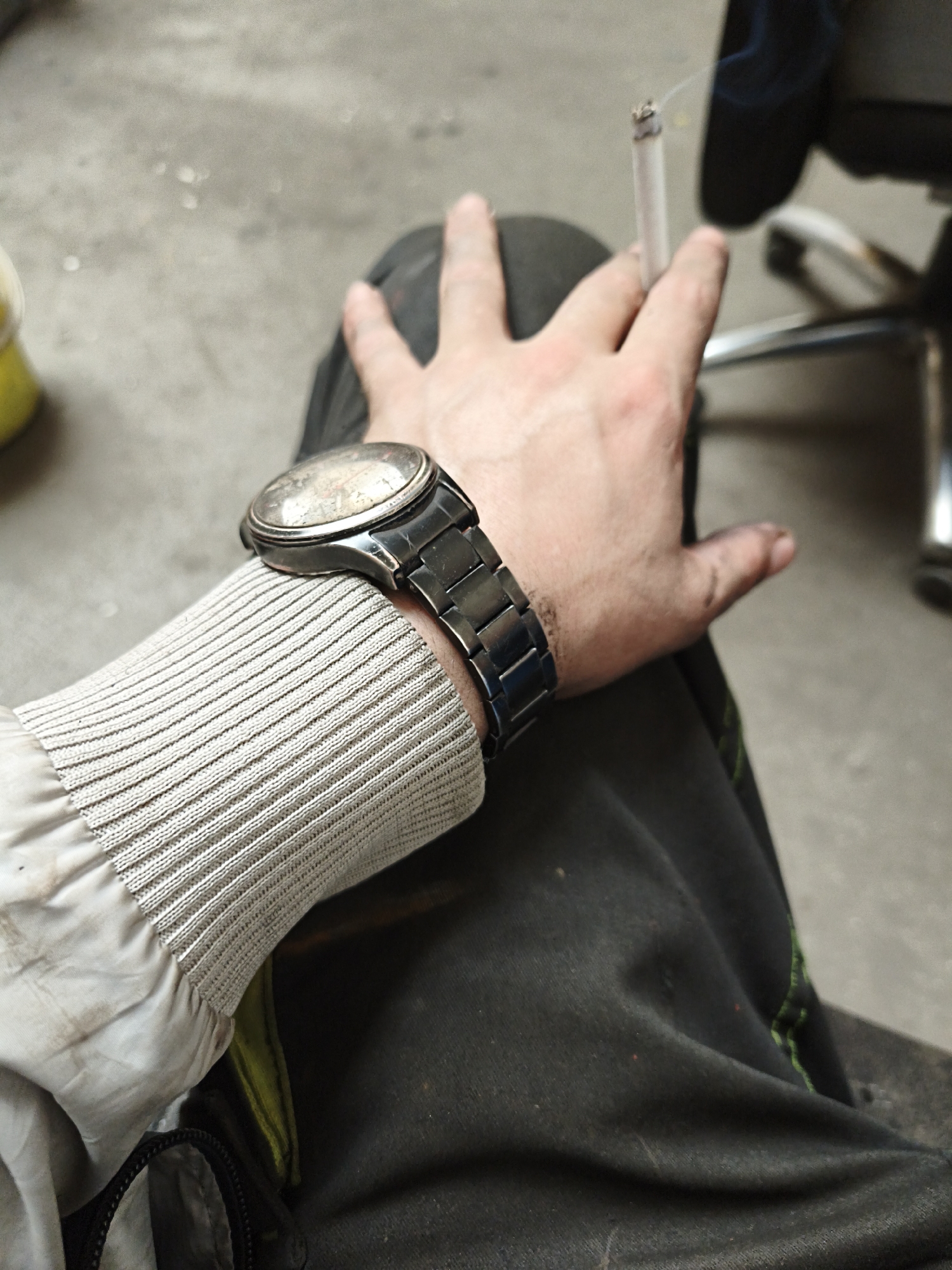 A close-up shot focuses on a person's right arm, adorned with a metallic wristwatch and clad in a grey, ribbed-cuff jacket. The person is holding a lit cigarette between their fingers, with a wisp of smoke rising upwards. Dark pants cover the lower portion of the arm, and a portion of a black bag with a zipper is visible. The background consists of a concrete floor and a metallic chair. The scene appears to be outdoors, potentially in Volevčice, Czechia, given the concrete ground and industrial-style chair.