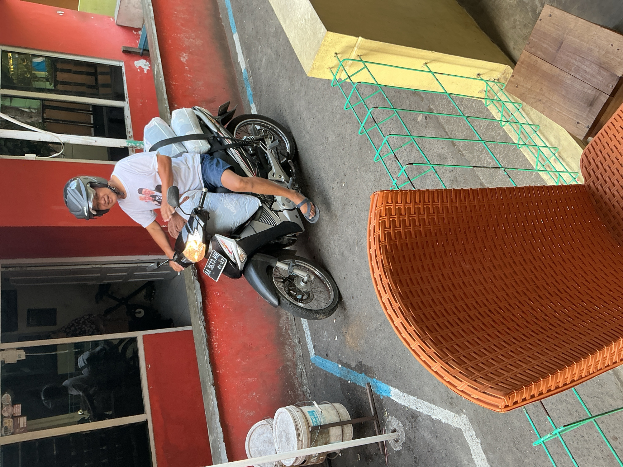 A man wearing a helmet and a white t-shirt with two portraits on it, blue shorts, and gray flip-flops is sitting on a motorcycle. The motorcycle has a license plate that reads "AA 5633 MH 01-27". The man appears to be looking at the camera and smiling. He has two white bags strapped to the back of the motorcycle. The motorcycle is parked in front of a red building with windows. In the foreground, there is an orange woven chair and a wooden bench. The scene appears to be outdoors, possibly in a residential area or a street in Magelang, Indonesia.