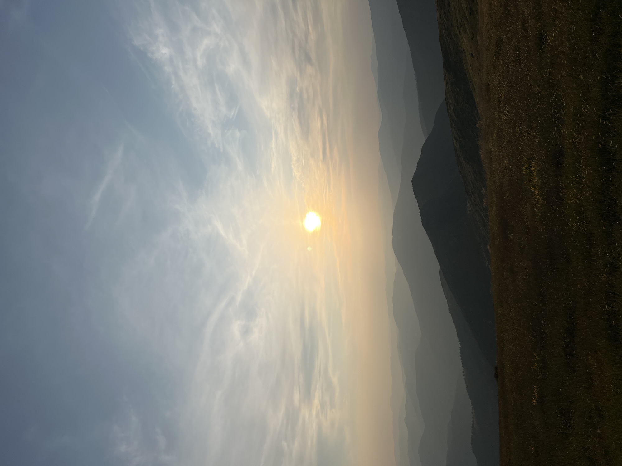 This image captures a serene mountain landscape at sunset, possibly near Luhy, Ukraine. The sun, a bright orb, is low in the sky, casting a warm, golden glow. Wispy clouds streak across the pale blue sky, tinged with orange and pink near the horizon. A series of mountain ranges recede into the distance, their silhouettes fading from dark gray to a soft, hazy blue due to atmospheric perspective. The foreground is dominated by a grassy meadow, with blades of grass catching the last rays of sunlight, appearing golden and brown. The overall impression is one of tranquility and natural beauty, with no people or man-made objects visible in the frame.