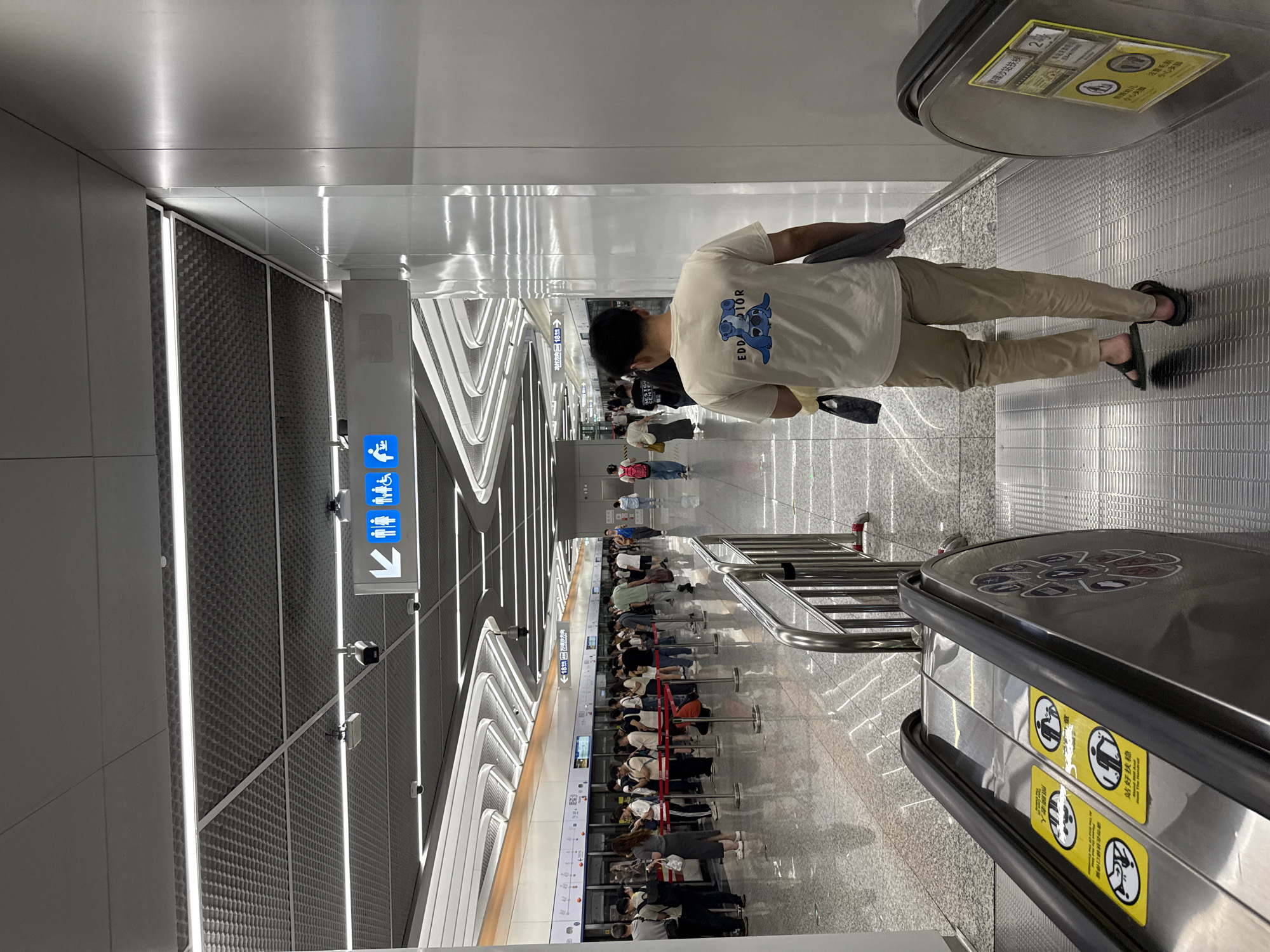 The image depicts an indoor scene in a subway station, presumably in Haizhu District, China, based on the presence of Chinese characters on the signs. A man stands on an escalator, wearing a white t-shirt with a graphic on the back, khaki pants, and sandals. He holds what appears to be a tablet or thin laptop. The escalator has signs in both Chinese and English, cautioning riders to stand and hold on.
The station features a long queue of people waiting near what appears to be a train platform. A sign indicates "冼村方向18號→", which could represent a train direction and platform number. Overhead lighting creates a striped effect on the ceiling. The station displays signage with symbols for accessibility (wheelchair, family, etc.).