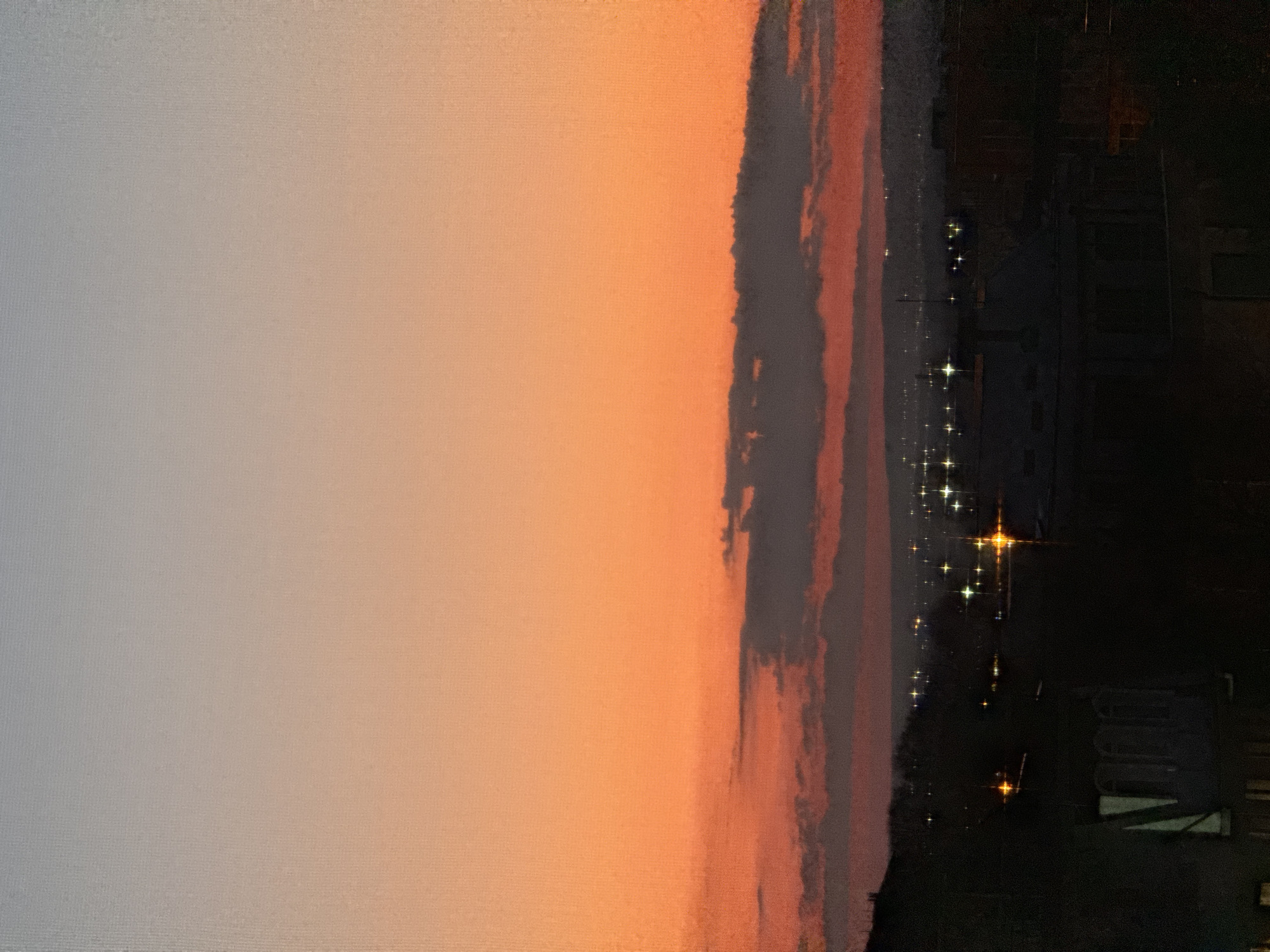 The image shows a cityscape at dusk or dawn. On the left is a gradient sky transitioning from light gray to orange. Further right, a band of clouds with gray and red hues is visible. The right side of the image features silhouettes of buildings and city lights, which appear as bright points of light. The location is Varna, Bulgaria, determined by the building silhouettes and characteristic layout of the cityscape. There are no people visible in the image. The scene depicts the transition from daylight to darkness in an urban environment.