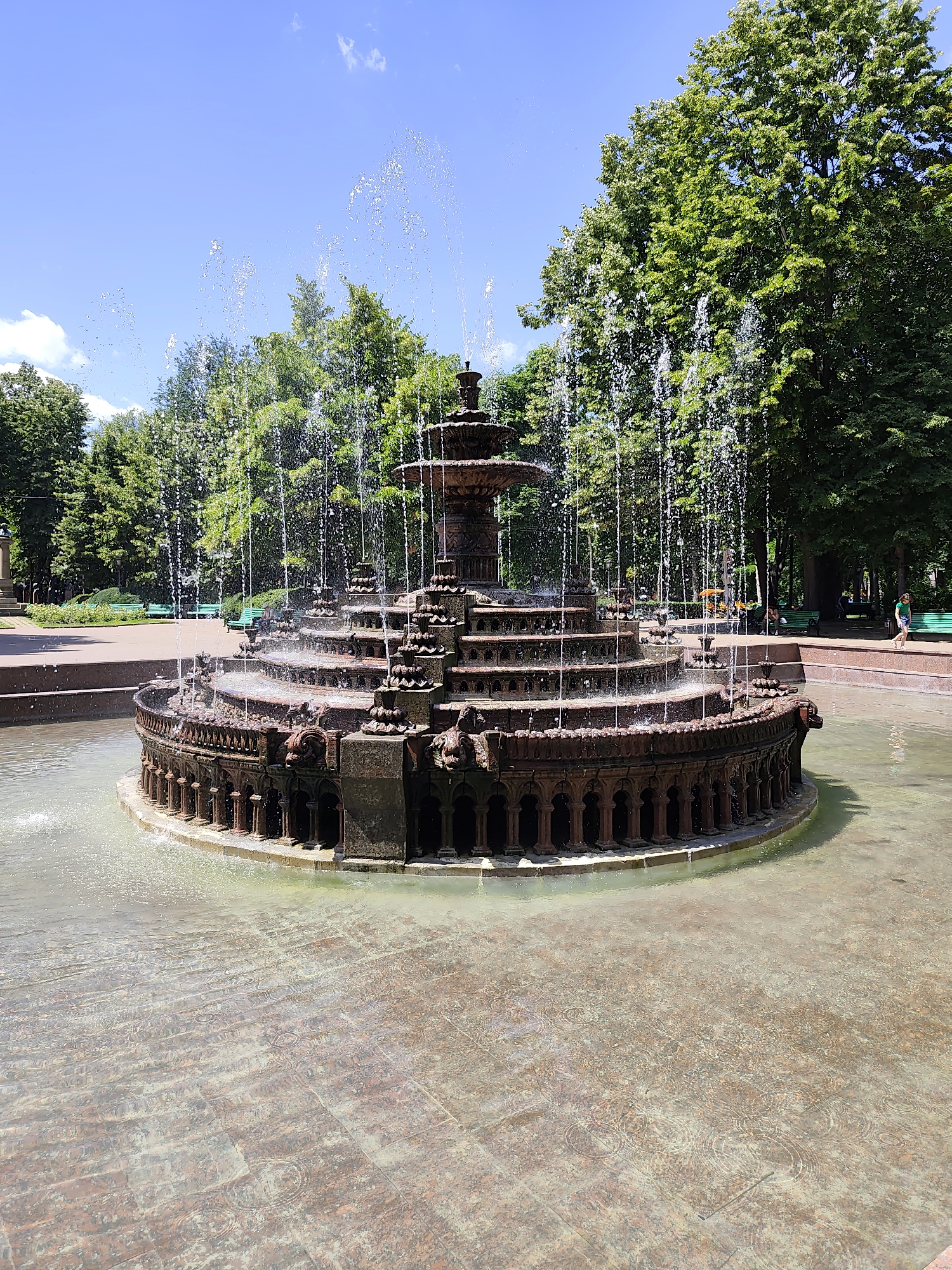 The image shows a large, ornate fountain with multiple tiers in a park setting. The fountain is made of dark, weathered material and features intricate carvings and architectural elements, including small columns and what appear to be animalistic faces. Water sprays from the top of the fountain and cascades down its various levels, creating a shimmering effect. The fountain is situated in a shallow pool with a patterned stone bottom that is visible through the water. Surrounding the fountain is a paved area and lush green trees. In the background, there are park benches and several people can be seen walking or relaxing in the distance. The sky is a clear blue with a few scattered white clouds. This fountain is located in Stephen the Great Central Park in Chișinău, Moldova.