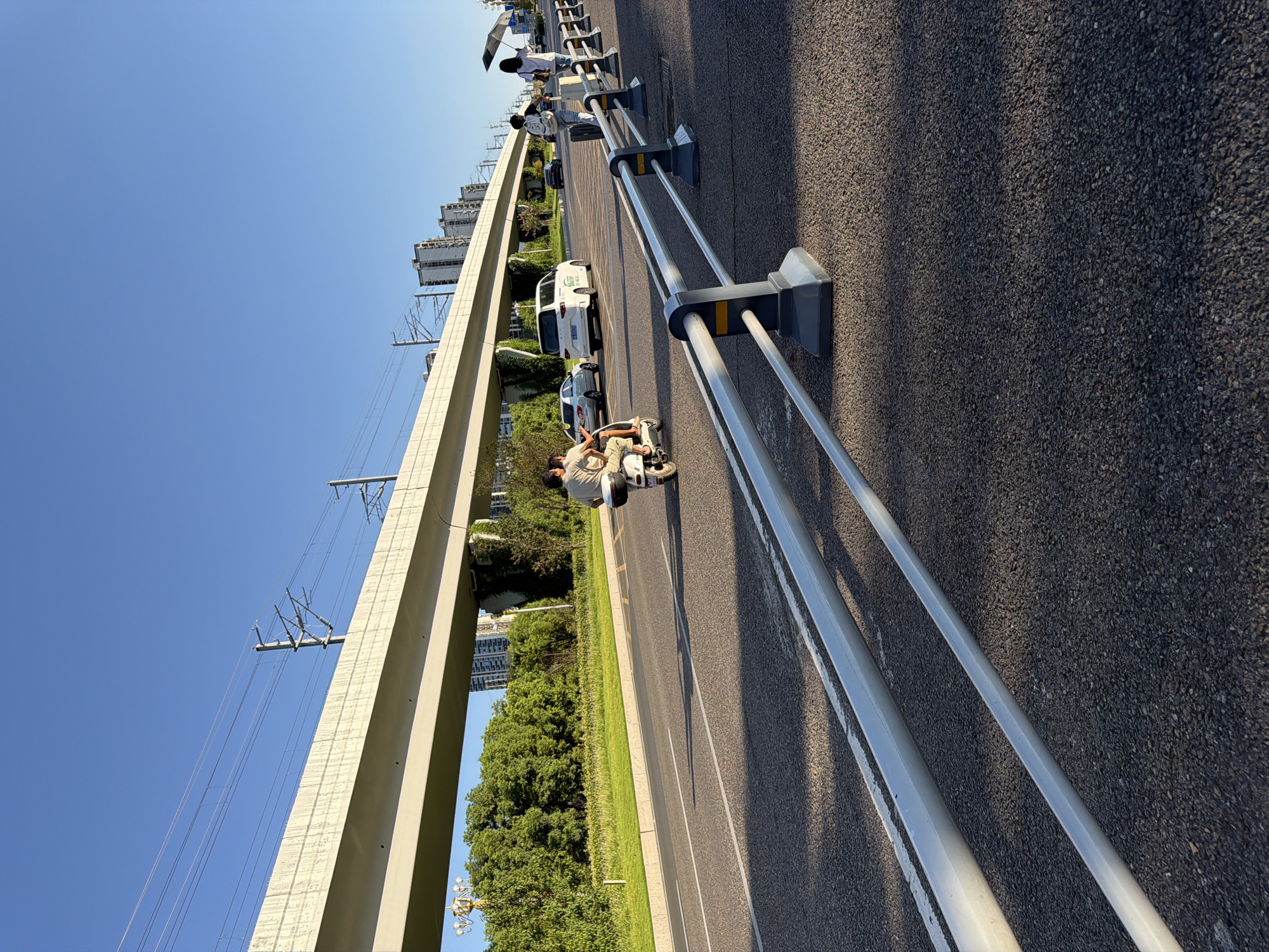 A sunny, clear day in Jiangjin, China, is depicted on a multi-lane road. An elevated structure, likely a highway or train line, runs horizontally across the upper portion of the frame, supported by concrete pillars and featuring overhead electrical lines. The road surface is asphalt with white lane markings. In the foreground, a silver metal barrier with dark grey supports separates the road from a sidewalk or pedestrian area.
On the road, a person rides a scooter with a passenger behind them. Further down the road, a white SUV is visible, followed by a black car. On the sidewalk, two individuals are walking. One is pulling a suitcase, and the other is holding a black umbrella. Lush green trees and vegetation line the side of the road, with tall buildings in the background. The light suggests it is late afternoon, casting long shadows.