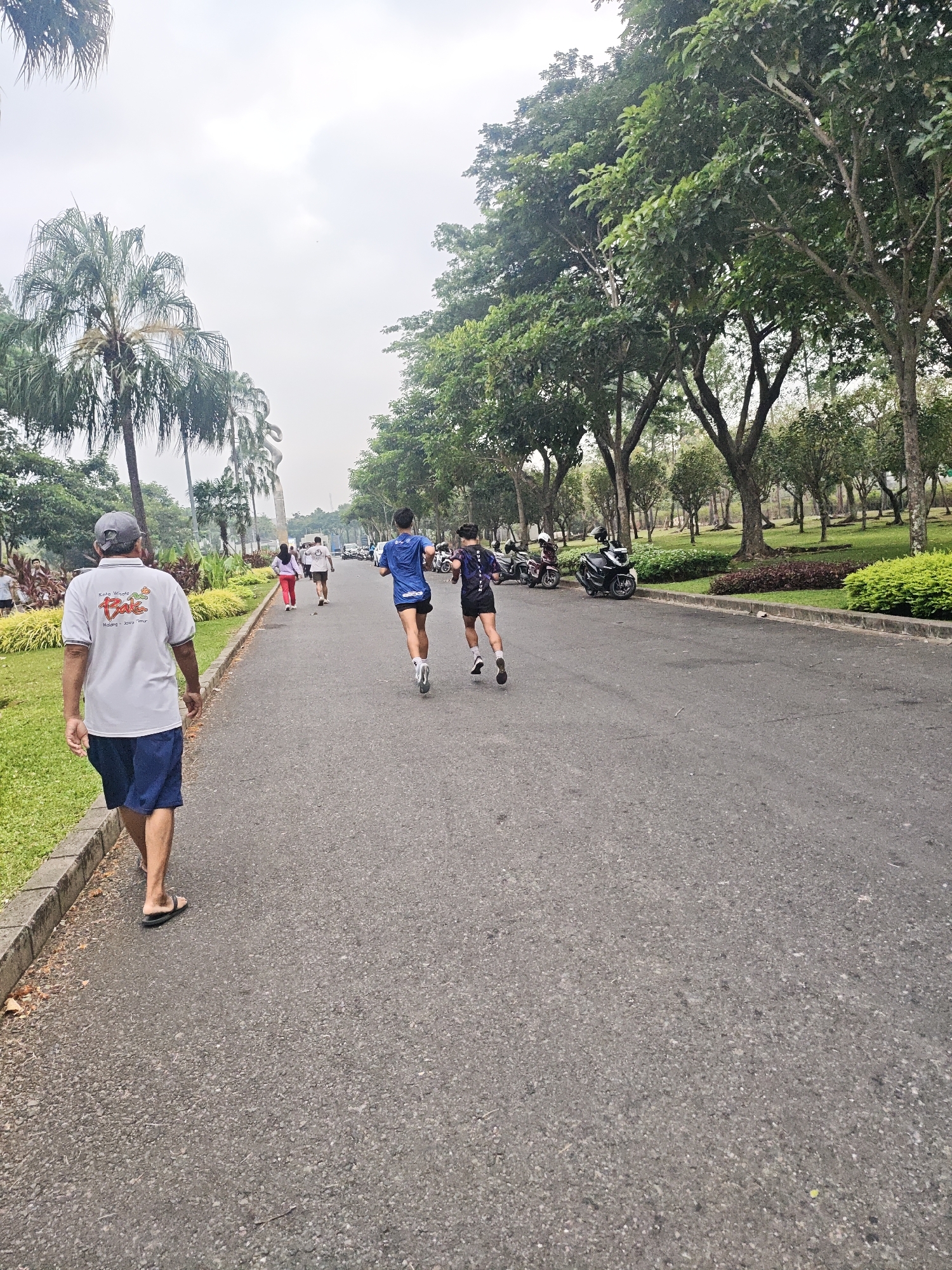 In Bacin, Indonesia, a man in a white t-shirt and blue shorts walks on an asphalt road next to a grassy area and palm trees. Two young men are running on the road ahead of him. In the background, there are more trees, some parked motorcycles, and a few other people walking. The sky is overcast.