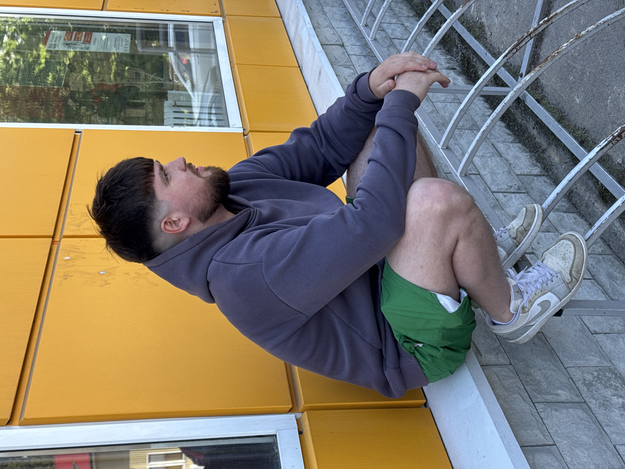 A man is depicted sitting outdoors, captured in a portrait-style shot that has been rotated 90 degrees counter-clockwise. He has dark, short hair styled with a fringe and a beard. He is wearing a dark gray hooded sweatshirt, bright green athletic shorts, and light-colored sneakers that appear worn. His hands are clasped together, resting on his knees, and he is gazing upwards and slightly to his right with a contemplative or observant expression.
The man is positioned on a low ledge or platform. To his left is a modern building with a distinctive bright yellow, panelled facade. A large window in this facade reflects the surroundings, including lush green trees and what appear to be other structures. Visible within the window's reflection are several signs or posters. One clearly shows the year "2024" and some partially obscured Cyrillic text, possibly related to work or employment. Another sign features a red background with white Cyrillic text, which could be "АКЦИЯ" (promotion/sale) or "УРА" (hooray), suggesting a commercial or celebratory context.
To the man's right, the ground is paved with grey tiles, and a low, curved metal railing, showing some signs of rust, runs alongside. The lighting suggests it is daytime, with clear weather. The scene offers a glimpse into a moment of rest or observation in Sosnovo, Russia.