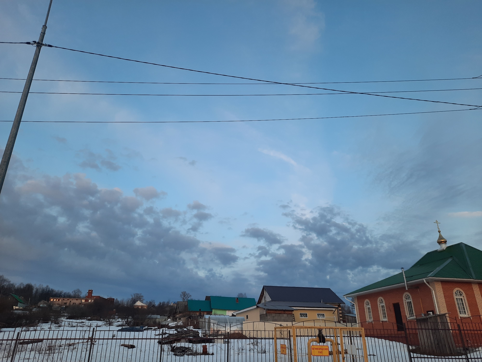 The image depicts a winter scene in what appears to be a residential area of Krasnogorskoye, Russia. The sky is overcast with patches of blue. Electrical wires stretch across the top half of the image. In the foreground is a snow-covered yard with a metal fence. Behind the fence are several residential buildings with various roof colors (green, gray). To the right is a red brick building, likely a church, with a green roof and a golden dome topped with a cross. A yellow gas pipeline runs along the fence. In the distance, behind the trees, more buildings are visible, including what appears to be a large brick structure.