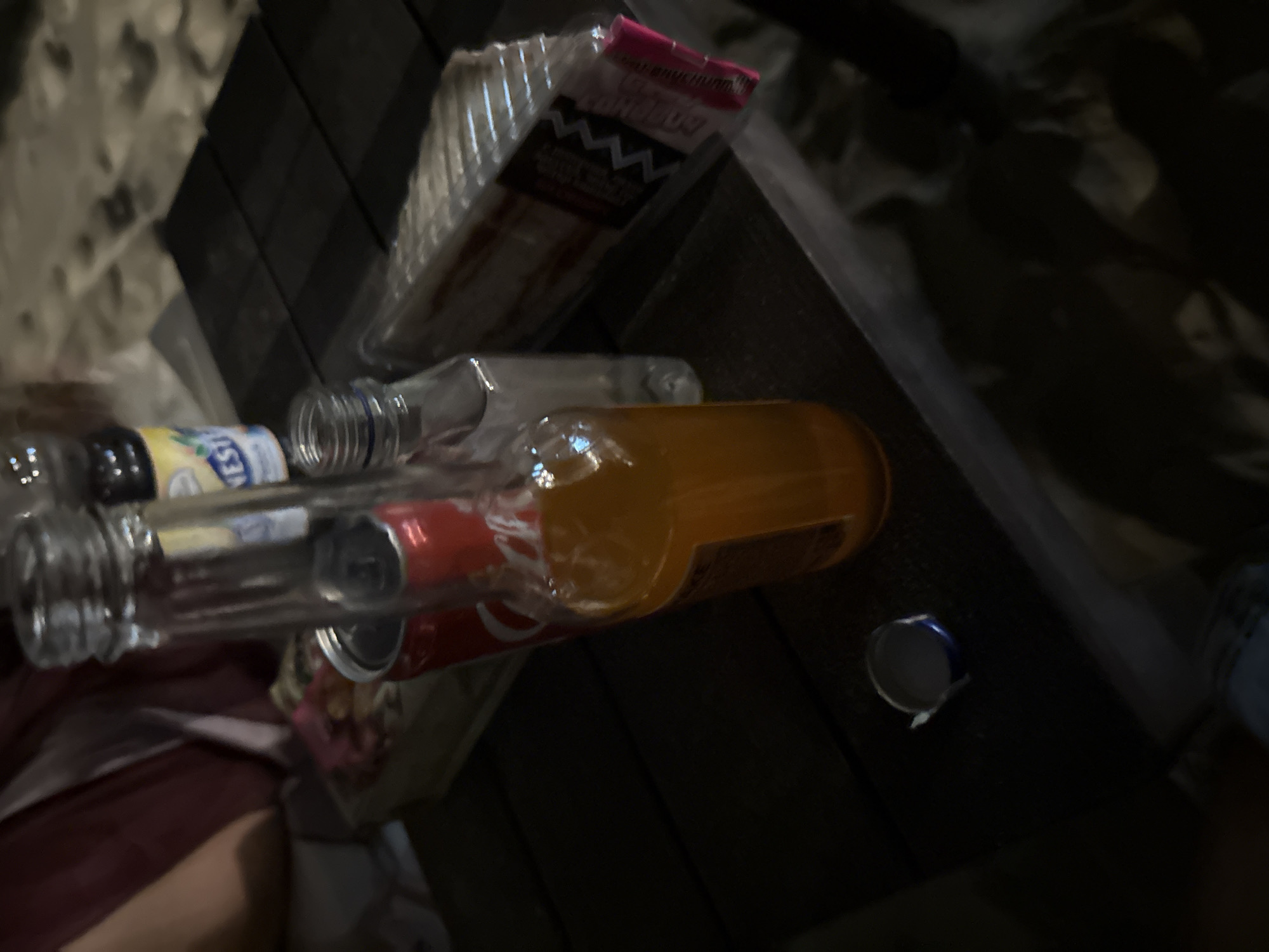 On a dark, wooden table surface, the image captures an assortment of items, including two beverage bottles, a can of soda, a packaged sandwich, and a detached can lid. In the foreground, a glass bottle is partially filled with an orange liquid, and next to it, a smaller plastic bottle is positioned. A red can, identified as Coca-Cola, is visible to the left of the orange drink. A packaged sandwich with visible layers of filling is placed to the right of the plastic bottle. A blue and silver can lid rests on the table near the bottom left corner of the image. The background is indistinct, but suggests a dim, possibly outdoor setting. The overall impression is that of a casual gathering or picnic. The location is identified as Sveti Vlas, Bulgaria.