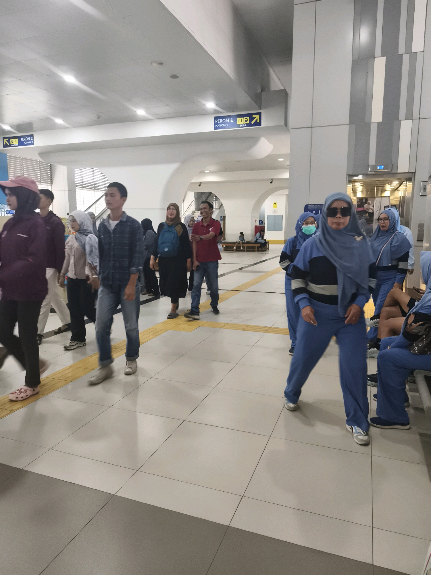 The image depicts a group of people walking through a modern, well-lit indoor space, likely a train station or transit hub, in Kelurahan 16 Ilir, Kec. Ilir Timur I, Indonesia. The environment features a tiled floor with a tactile warning strip, indicative of a public transportation area. Overhead, fluorescent lights illuminate the space, and architectural elements such as columns and signage are visible.
The individuals in the foreground and midground are dressed in a mix of casual and more uniform attire. Several women are wearing headscarves (hijab), with some in blue uniforms and others in darker, more casual clothing. One woman in the mid-right is wearing dark sunglasses and a blue headscarf and blue tracksuit. Men are also present, one in a blue plaid shirt and jeans, and another in a red polo shirt and jeans. The people are walking in various directions, suggesting movement and transit. Some individuals appear to be interacting, while others are walking independently. In the background, an elevator with visible passengers inside, and directional signs pointing to "PERON 1" (Platform 1) and "PERON 2" (Platform 2), further confirm the location as a transportation terminal. The overall impression is one of a bustling public space during daylight hours.