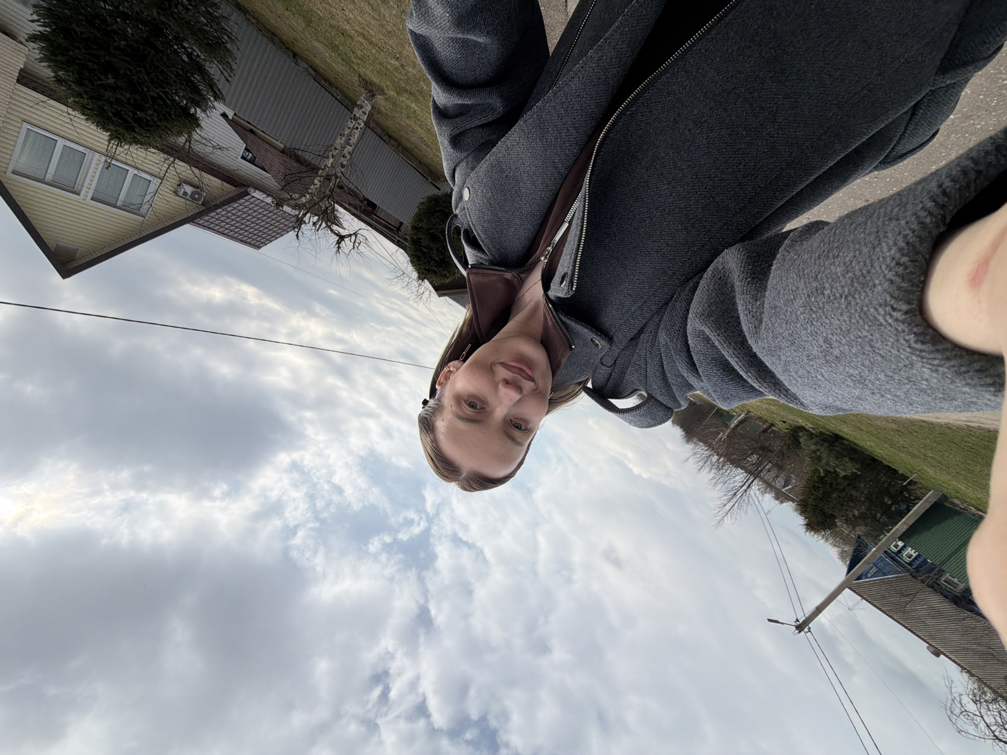 A selfie photograph of a fair-skinned woman is oriented with the sky and cloudy horizon at the bottom of the frame. She is wearing a dark grey coat with a brown collar visible. To the left, a yellow house with a brown roof and white-framed windows is visible, along with a small green bush. In the background, power lines cross the sky. To the right, a partially visible green and blue building with a corrugated roof stands amidst a grassy area and leafless trees. The image was taken outside in Reshetnikovo, Russia.