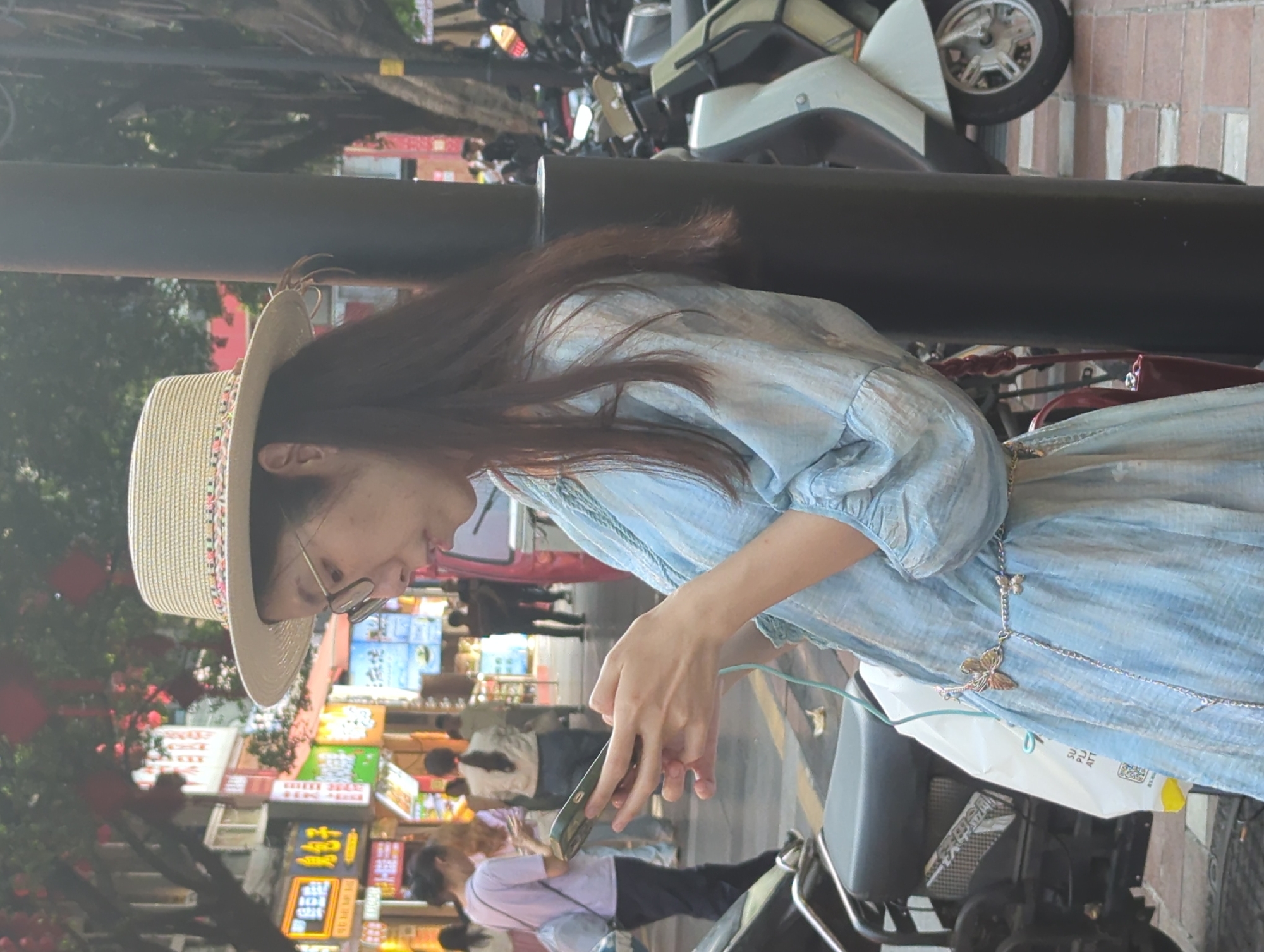 A young woman is captured in a side profile on a bustling street, looking down intently at her smartphone. She is wearing a light blue, flowing dress with puffed sleeves and a wide-brimmed straw hat adorned with a colorful band. Her long, dark hair cascades over her shoulder, and she wears eyeglasses. Around her waist, a delicate silver chain with two butterfly ornaments is visible. She holds the smartphone, which has a light green case, with both hands.
The background reveals a vibrant outdoor urban setting in Yuzhong District, China, under bright daylight. Numerous shops line the street, featuring colorful signs with Chinese characters, some of which are illuminated. Other pedestrians, some blurred, are visible walking and standing. Trees with red decorations, possibly lanterns or blossoms, add a festive touch to the left. Parked and passing scooters or motorcycles are present throughout the scene, with parts of one scooter clearly visible on the right. A prominent black pole stands in the mid-ground. A white plastic bag with some indistinct text hangs from a vehicle or railing near the woman. The overall atmosphere is one of a busy commercial area.