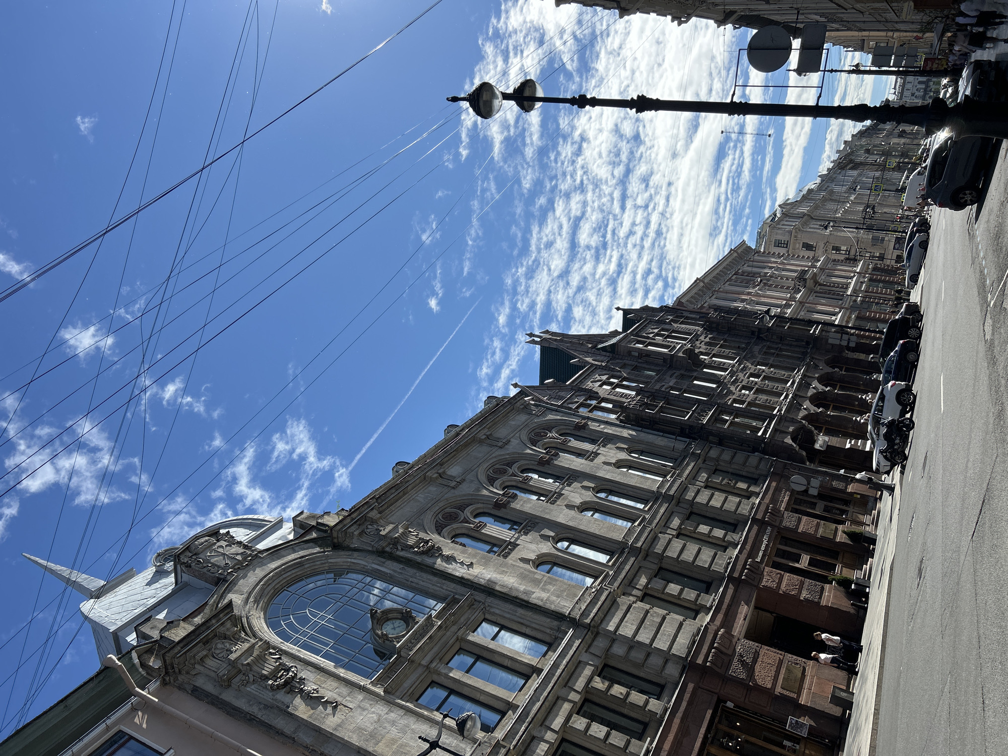 This is a tilted, eye-level shot of a street scene in Unknown, Unknown, featuring ornate, aged architecture and a bright blue sky filled with wispy clouds and electrical wires. A grand, stone building with multiple arched windows, decorative carvings, and a prominent clock face dominates the left side of the frame. The building's facade is a mix of grey stone and darker reddish-brown lower levels. A tall, ornate lamppost with two lights stands on the right side of the street. The street itself is paved and shows car traffic, including parked cars, a motorcycle, and vehicles in motion. Sidewalks are visible along the buildings, with a few pedestrians walking. The overall impression is of an established urban environment with a mix of historical architecture and modern life.