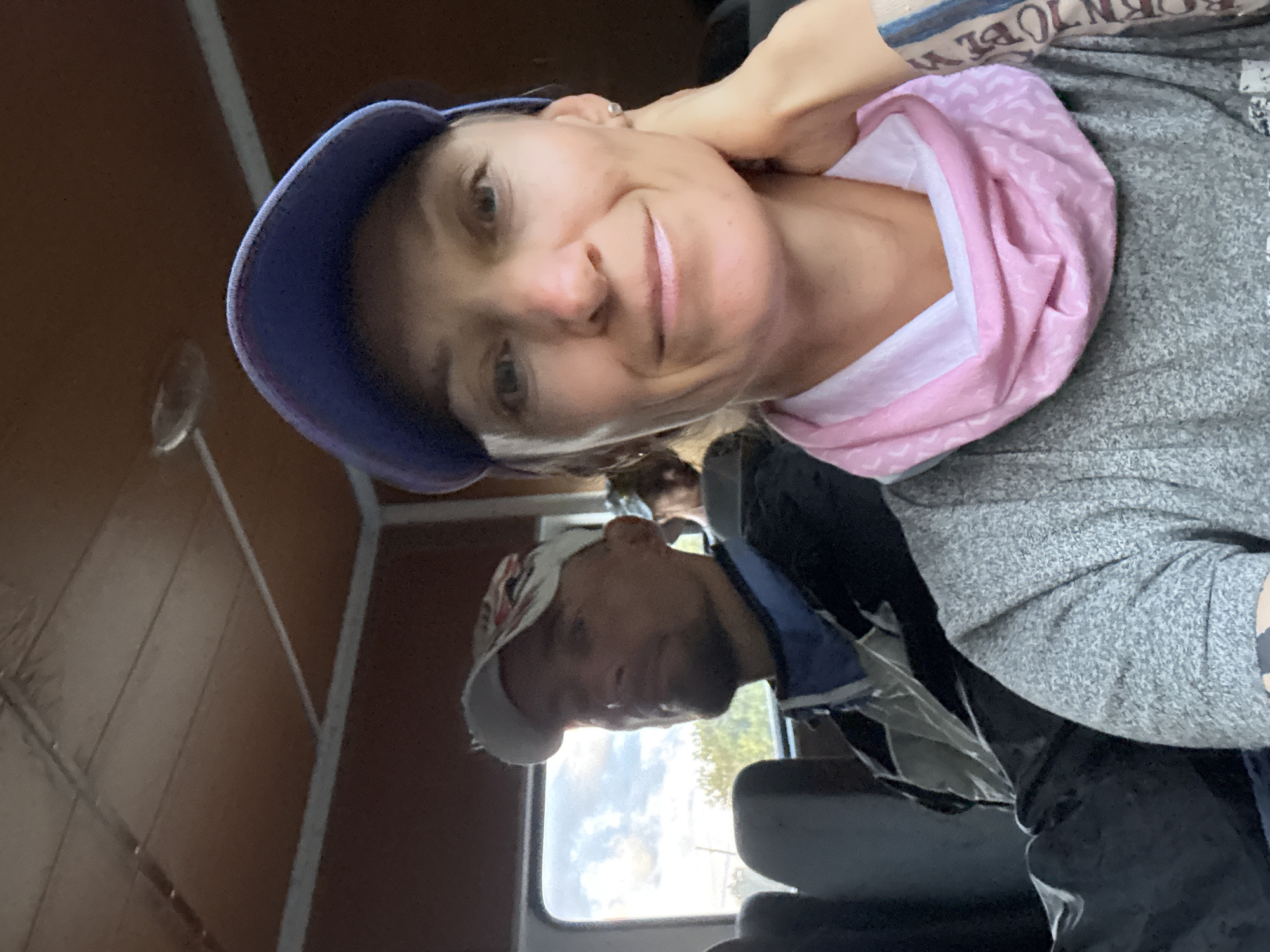 This is a selfie taken from inside a vehicle. The foreground features a woman wearing a blue baseball cap and a gray long-sleeved shirt with a pink and white neck gaiter. She has her right hand resting on her cheek. Behind her and to her left, a man wearing a white baseball cap and a blue and gray jacket is visible, looking towards the left. The interior of the vehicle appears to have a wood-paneled ceiling. Outside the window, trees and a cloudy sky are visible. The overall setting suggests a travel or transportation context, possibly within the city of Lavrovoe, Ukraine, though the image itself does not provide explicit confirmation of this location.