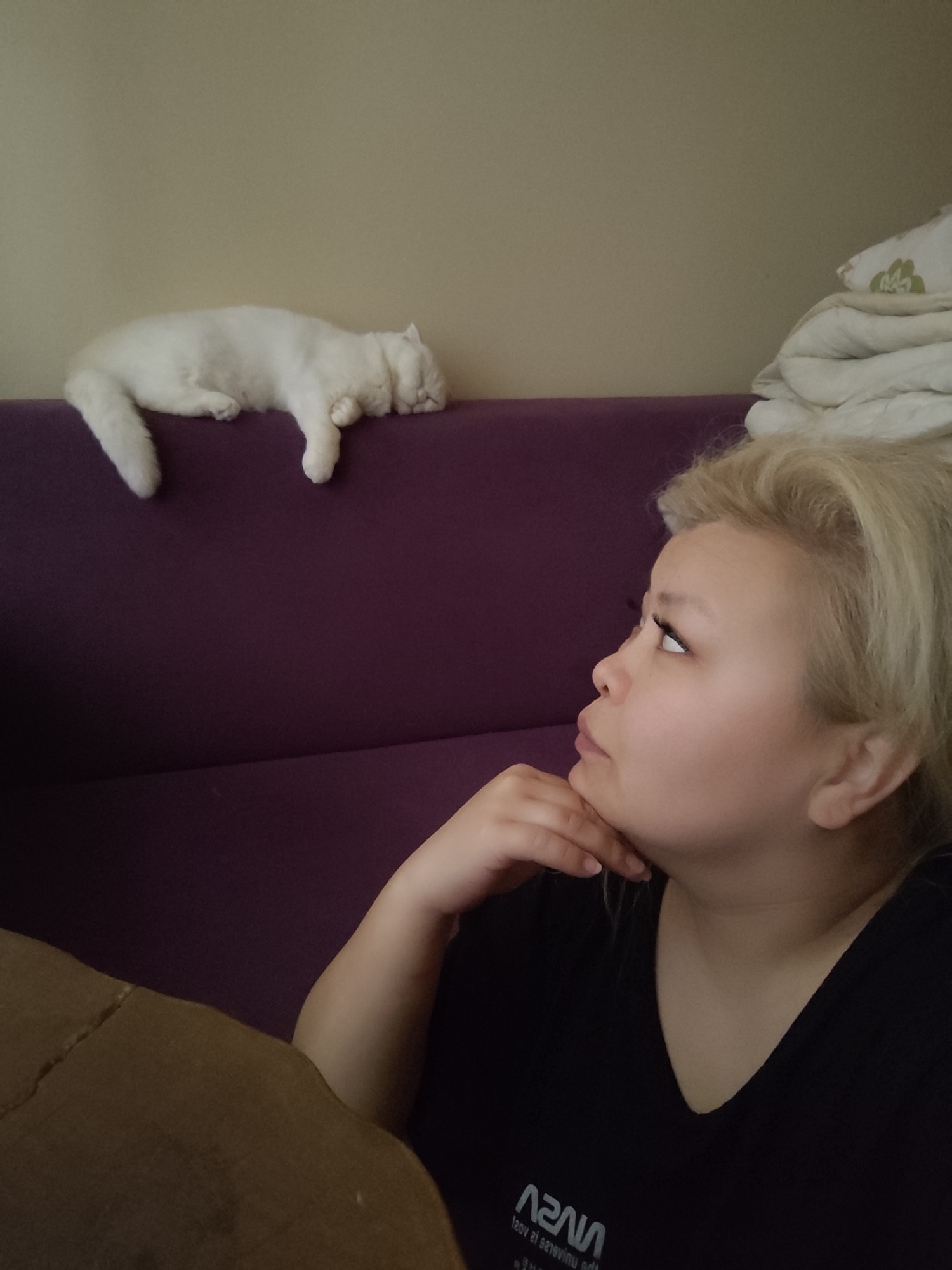 A medium shot captures a person and a cat in an indoor setting that seems to be a home. The person, who appears to be a woman, is on the right side of the frame, looking upwards and slightly to the left with her hand resting on her chin as if deep in thought. She has blonde hair and is wearing a black t-shirt with white text that is a mirrored image. To the left and in the upper part of the frame, a white cat is sleeping on top of a purple sofa. The background is a light-colored wall. The lower left corner of the image shows a portion of what appears to be a wooden table. There is no clear information indicating that the image was taken in Sarıyer, Turkey.
