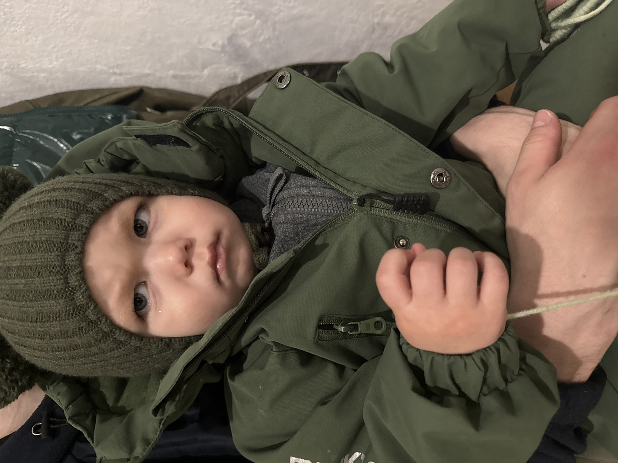 A young child, likely a baby or toddler, is depicted in a close-up, vertically oriented shot. The child is dressed in warm winter attire, featuring an olive green knitted hat, possibly with ear flaps, and a thick olive green hooded jacket with a zipper and snap fasteners. A grey zipped top is visible underneath the jacket.
The child's blue eyes gaze directly towards the viewer with a neutral, somewhat pensive expression. Their small right hand is curled around a light green string or piece of yarn. An adult's arm and hand are visible on the right side of the frame, appearing to support or hold the child's leg.
The setting appears to be indoors, with a textured white wall visible in the upper background. Other dark green and blue fabrics, possibly blankets or additional clothing, are present in the background and underneath the child. The warm clothing suggests cold weather conditions, aligning with the location context of Chaikovsky, Russia. The lighting is soft and diffused, not indicating a specific time of day. Some white, indistinct text is partially visible on the lower right portion of the child's jacket.