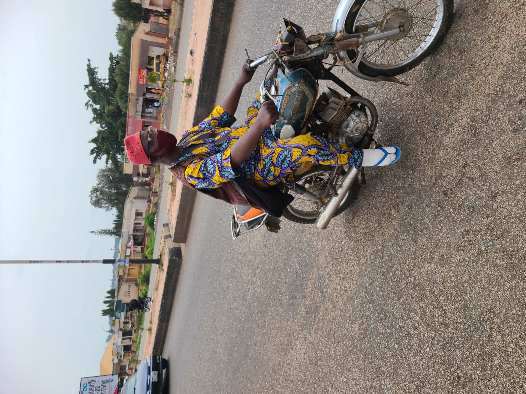 A man wearing a red hat, glasses, and a colorful yellow and blue patterned outfit is riding a motorcycle on a paved road. He is wearing white socks and blue flip-flops. The motorcycle appears old and somewhat rusted. The background shows a city street with buildings, trees, and utility poles. Based on the prompt, the location is Gura Top, Nigeria. A car is visible in the bottom left corner.