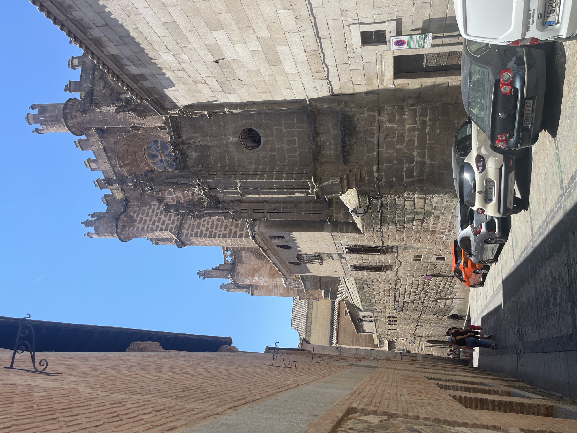 This image depicts a narrow cobblestone street in Toledo, Spain, with historic stone and brick buildings lining both sides. On the right, a large, imposing stone structure, likely a cathedral or church, dominates the scene with its intricate Gothic architecture, including arched windows, crenelated towers, and decorative stonework. Sunlight casts distinct shadows across the street and buildings. Several cars are parked along the right side of the street, including an orange hatchback, a silver sedan, a white SUV, and the rear of a white van. On the left, a brick building with an awning and metal brackets runs along the street. A small group of tourists are visible walking down the street towards the left, appearing to be exploring the historic area. The sky is a clear, bright blue.