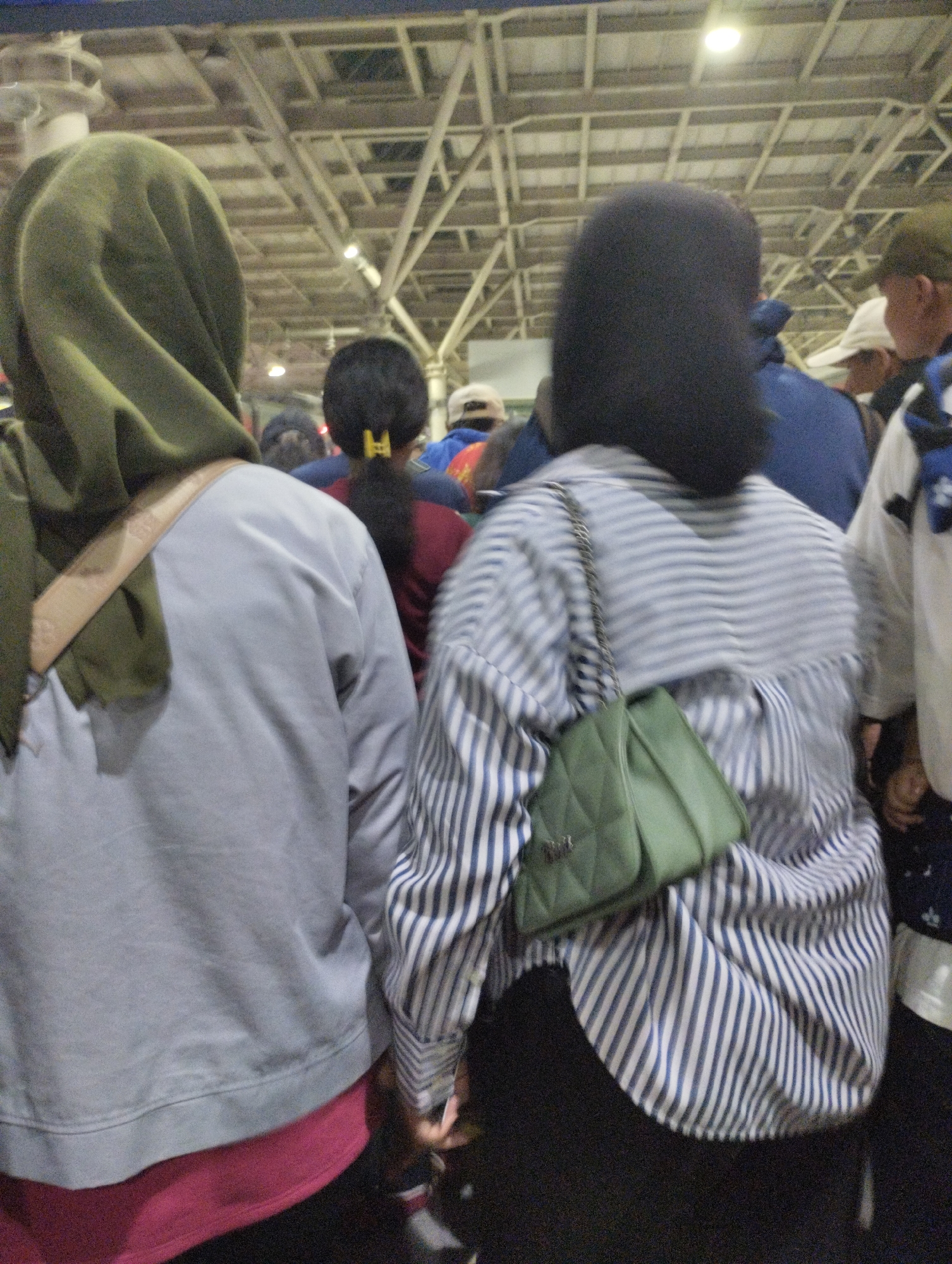 The image shows the back of several people standing in what appears to be an indoor market or exhibition hall, possibly at the Jakarta Convention Center in South Jakarta, Indonesia. The ceiling features exposed industrial beams and lighting. In the foreground, on the left, a person wearing a light grey sweatshirt and a green hijab is seen from the back, with a tan strap across their shoulder. To their right, another person is visible from the back, wearing a blue jacket and a red shirt underneath, with their dark hair pulled back with a yellow clip. In the center-right, a person in a striped blue and white shirt carries a green quilted handbag on their shoulder. Other individuals and clothing items are blurred in the background. The lighting suggests an indoor setting, likely during the day.