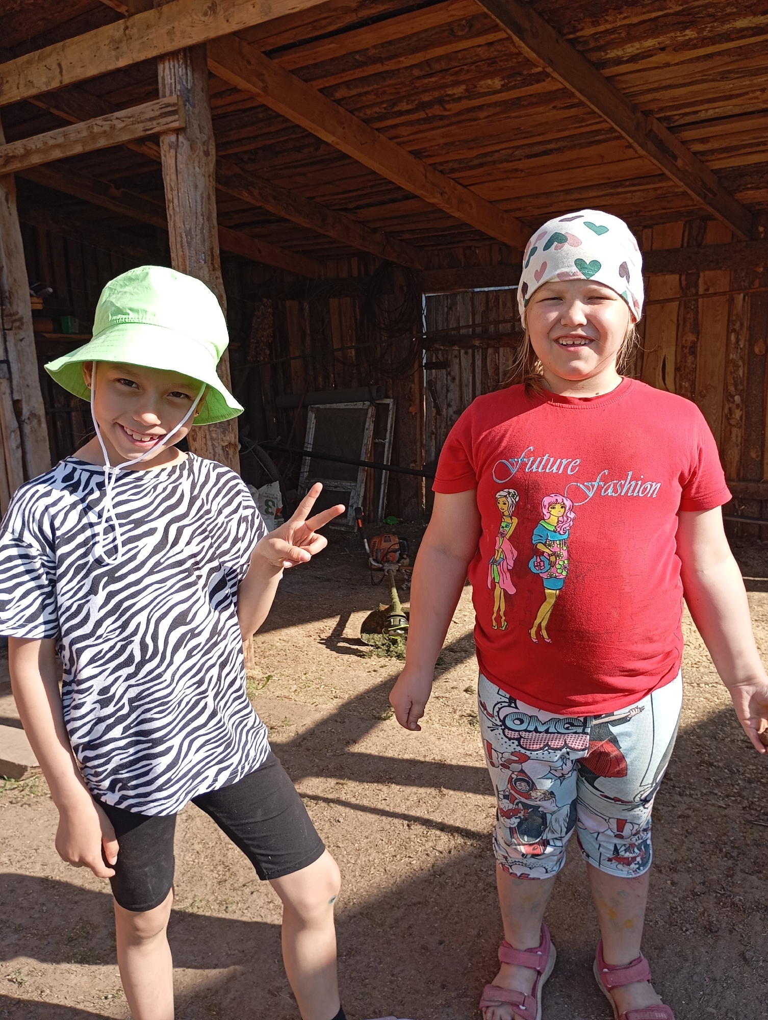 Two smiling young girls stand in front of a wooden structure in an outdoor setting. The girl on the left wears a zebra-print shirt, black shorts, and a bright green hat, flashing a peace sign with her right hand. The girl on the right is dressed in a red T-shirt with a graphic design and comic-themed patterned leggings, complemented by pink sandals and a patterned head covering. Behind them is a rustic wooden shed or outbuilding, partially obscured by shadows, with a glimpse of tools or equipment inside. The ground appears to be dirt or gravel, and sunlight illuminates the scene. Location: Unknown, Unknown.