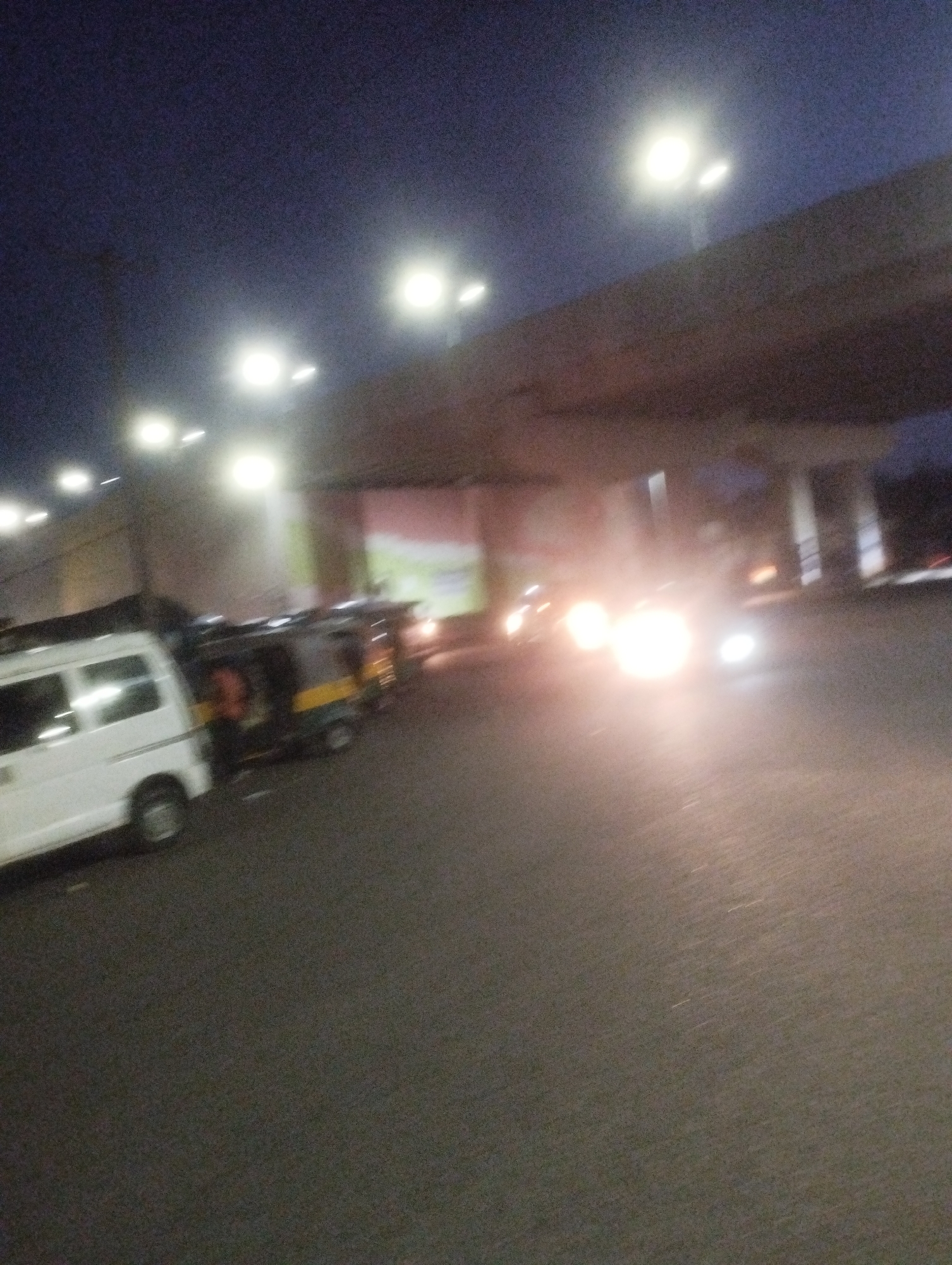 The image depicts a street scene at dusk or night in Jos, Nigeria, under an elevated road structure. A white van is parked on the left side of the road. To its right, several auto-rickshaws are also parked. Streetlights are visible in the background, casting a glow. The road surface is asphalt. The lighting suggests that the photo was taken during a low-light condition, possibly with the use of artificial light sources. The scene appears to be an urban environment, consistent with a city like Jos. There are no discernible individuals in the image.