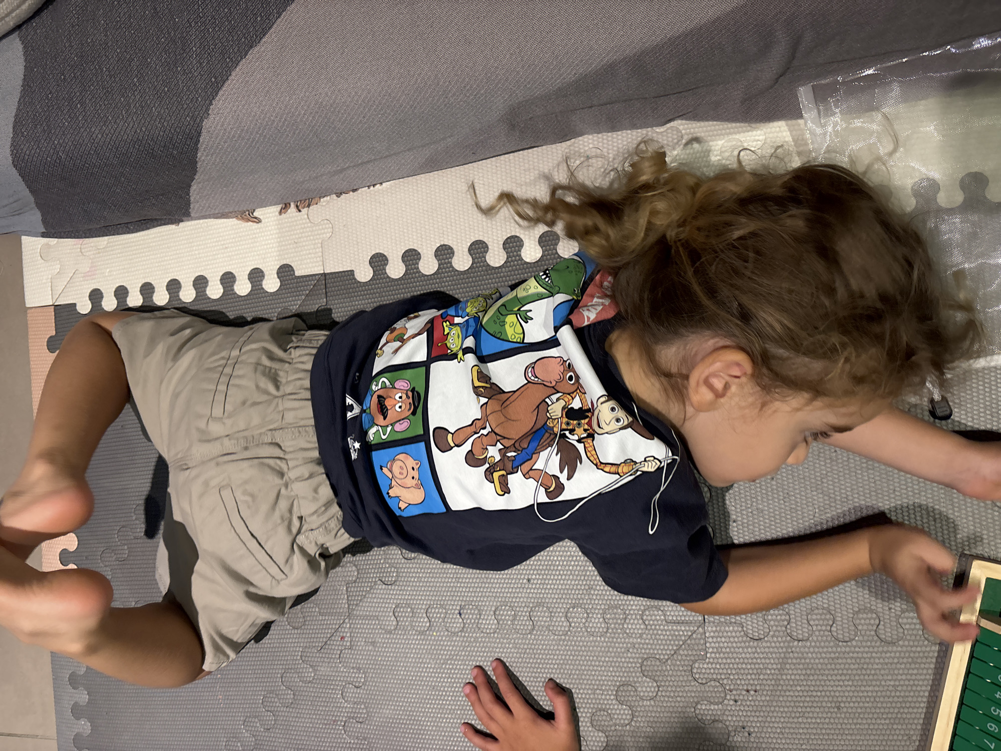 A young child lies on their back on a puzzle mat, upside down, with their feet in the air. The child is wearing a dark blue t-shirt with a graphic of characters from the movie Toy Story, including Woody, Bullseye, Hamm, Rex, and the Little Green Men. The child also wears light brown shorts. The background consists of a grey and white puzzle mat and a section of a grey upholstered couch. The scene is indoors. The child's hair is curly and light brown, and is visible on the right side of the image. A small hand with visible fingers is on the left side of the image. A wooden toy with green numbered tiles is visible in the bottom left corner. There is no visible indication of the city of Dubai, United Arab Emirates.