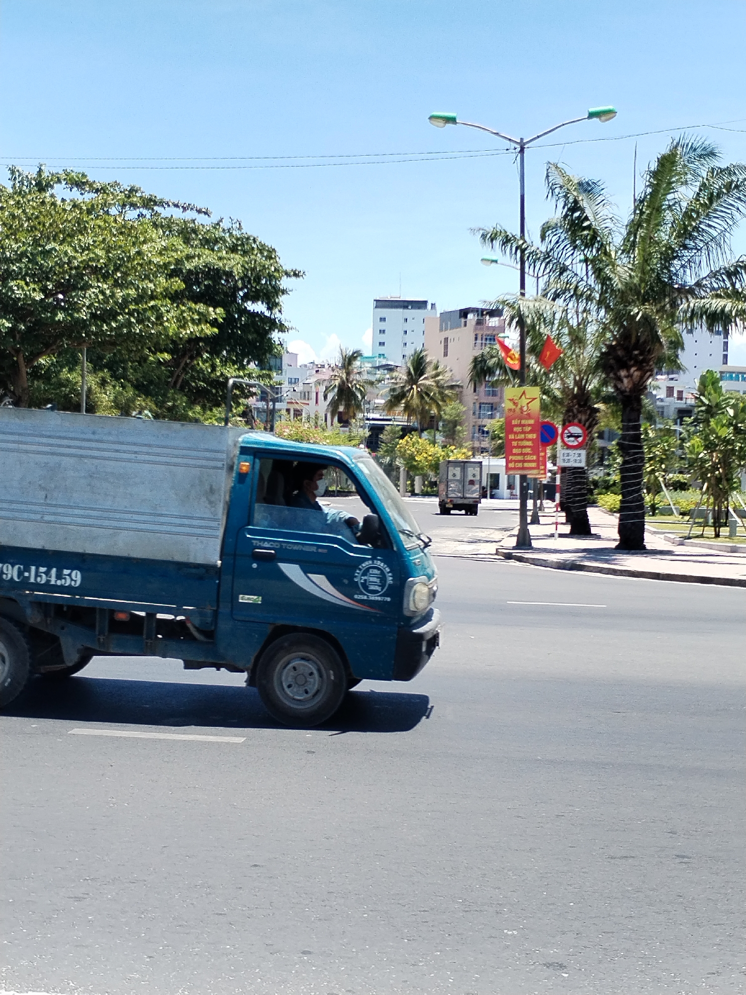 The image shows a blue Thaco Towner truck driving on a city street in Nha Trang Ward, Vietnam. The truck, with license plate 79C-154.59, has a silver-colored cargo bed. The driver is visible inside the cab, wearing a mask. In the background, there are buildings, palm trees, and a street light. A red and yellow sign with Vietnamese writing is visible on the side of the road, along with another sign restricting vehicle access between certain hours (6:30 - 7:30 and 16:30 - 18:30). The sky is blue, and the weather appears to be sunny.