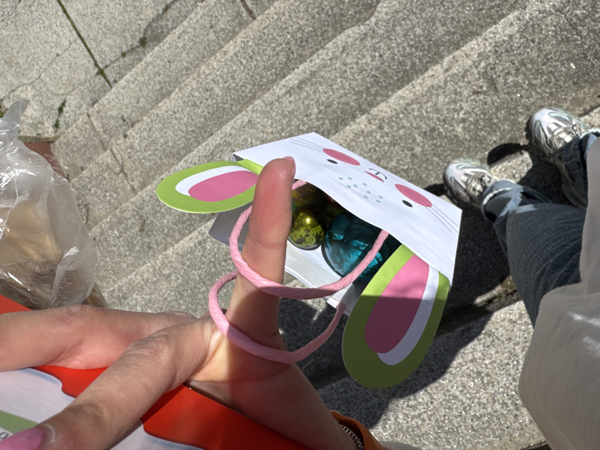 A person stands on concrete stairs, holding a white paper bag decorated with a bunny face and pink and green ears, filled with gold and teal objects that appear to be Easter eggs. The bag's pink twine handle is looped around a finger. A plastic bag of unidentified contents is visible to the left. The person is wearing jeans and silver sneakers. The location appears to be outdoors, possibly a public space with stone steps. The scene's location in Blagoevgrad, Bulgaria, cannot be determined.