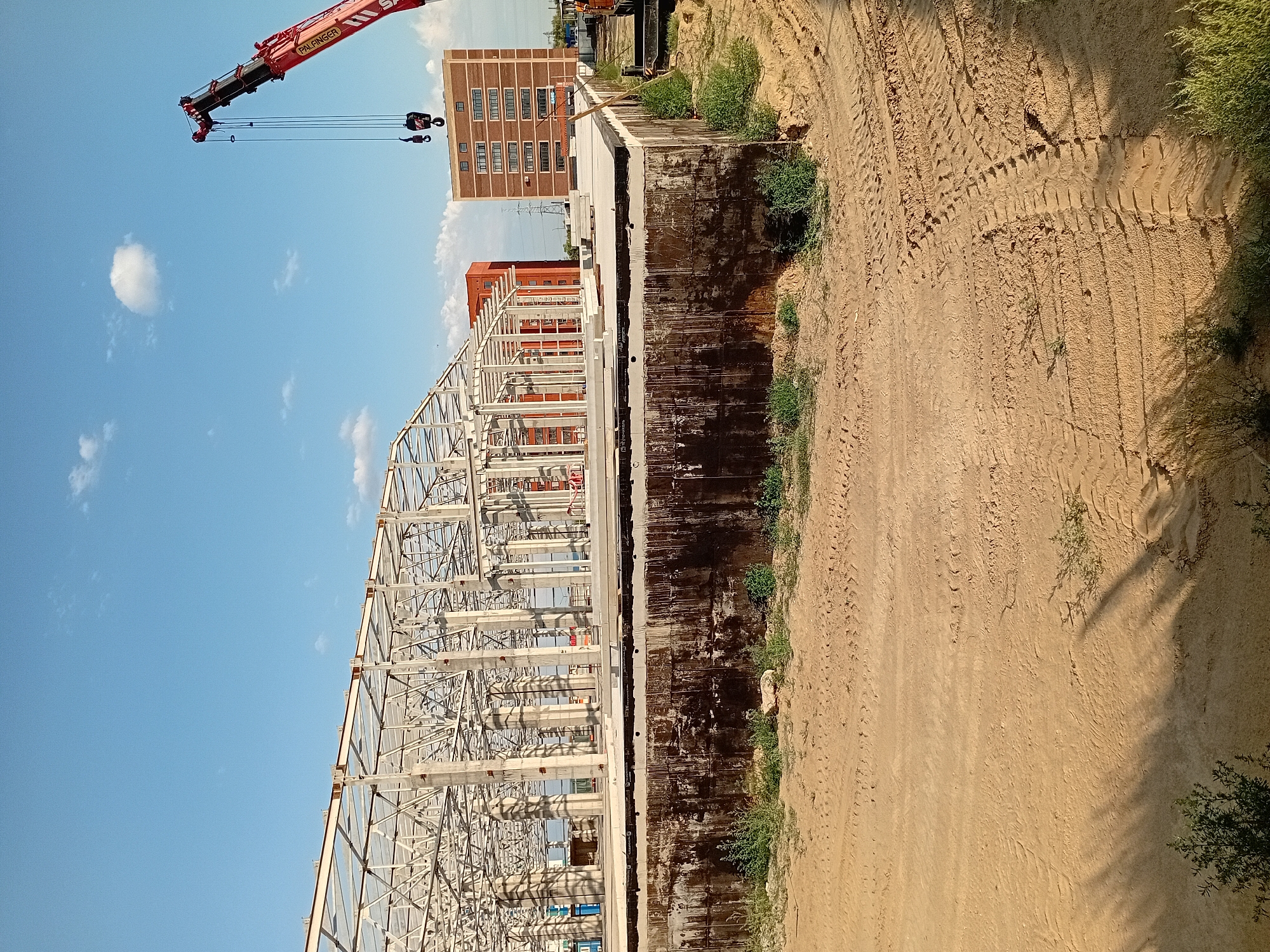 The image depicts a construction site in the city of Semey, Kazakhstan, under a clear blue sky with a few scattered white clouds. In the foreground, there is a large foundation structure, possibly for a building. The foundation is surrounded by sandy ground with visible tire tracks from heavy machinery.
Dominating the middle ground is the partially constructed steel frame of a large building. Numerous concrete columns support a complex network of steel beams and trusses, indicating the early stages of erecting a substantial structure. Behind the steel frame, two multi-story buildings are visible. One is a modern, red brick building with numerous windows, and another appears to be an orange-colored building.
In the upper right portion of the image, a red crane with its boom extended is positioned over the construction site, suggesting ongoing work and the presence of heavy lifting equipment. The overall scene conveys an active construction environment in progress.