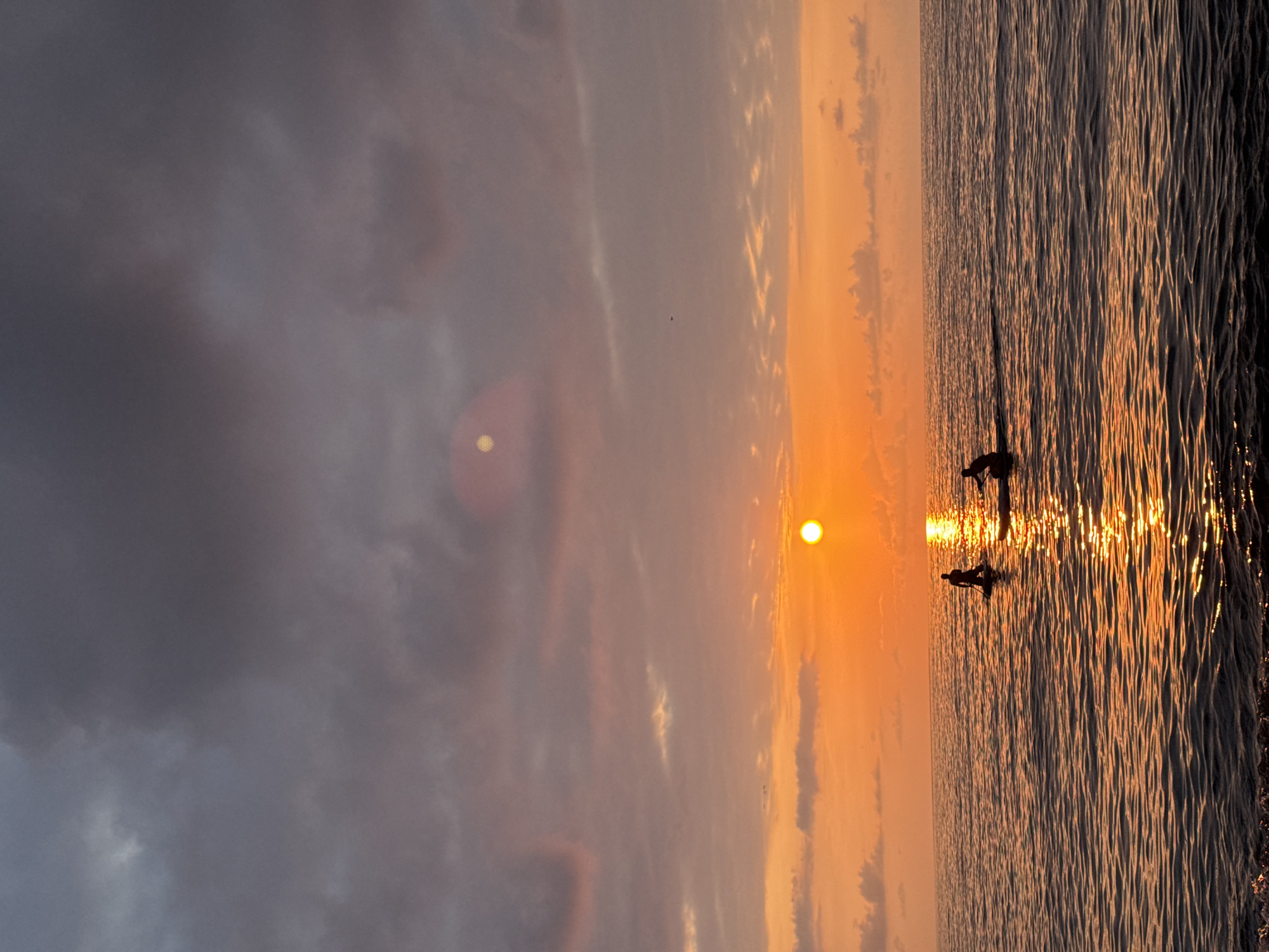 This image captures a serene sunset over the Black Sea, likely near Batumi, Georgia. Two silhouetted figures are visible on paddleboards in the water, seemingly enjoying the tranquil evening. The sun, a bright orb, is low on the horizon, casting a warm, golden glow across the water and illuminating the scene with vibrant orange and pink hues. The sky is a mix of dramatic clouds, with darker tones towards the top of the frame gradually transitioning to softer, lighter shades as they approach the horizon. The water exhibits gentle ripples, reflecting the light of the setting sun and creating a shimmering effect. In the foreground, the dark edge of a beach or shoreline is visible. The overall atmosphere is peaceful and picturesque, indicative of a relaxing moment at the coast.