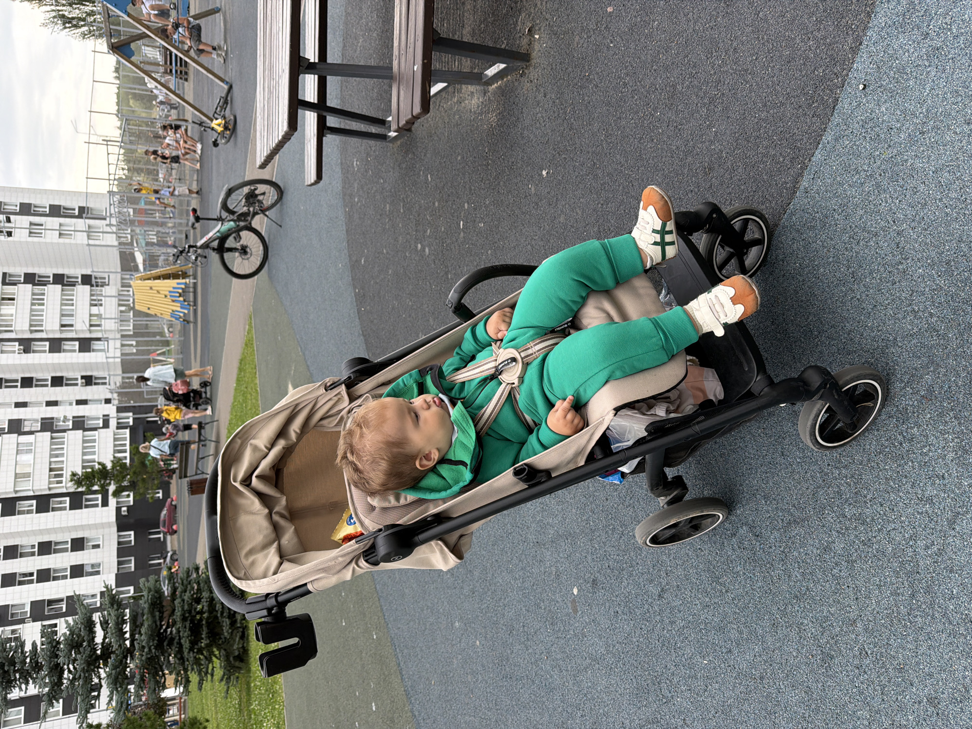 The image depicts a young child sitting in a stroller outdoors, likely in a public park or playground, with a modern apartment building in the background. The child is wearing a bright green tracksuit and white and orange sneakers. The stroller is a light beige color with black accents and wheels. In the background, there is playground equipment, including swings and climbing structures, as well as several bicycles. A few adults and children are visible in the distance, suggesting a family-friendly environment. The ground is paved with a dark grey and blue speckled material, and there is some green grass visible to the left. The overall atmosphere appears to be casual and relaxed, with the weather seeming overcast. While the image is not explicitly tagged with the city of Kazan, Russia, the architectural style of the apartment building is consistent with modern construction found in Russian cities.