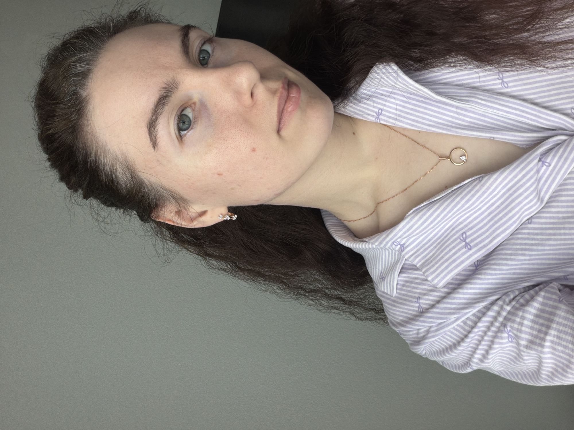 A woman with fair skin and dark brown hair is seen from the chest up, with the image rotated to the left. She has blue eyes and a neutral expression. She wears a lavender and white striped collared shirt with small bow designs and a delicate gold necklace with a circular pendant. Her left ear is visible, adorned with a small, intricate earring. The background is a plain gray surface. There is no evidence to suggest the location is Yanino-1, Russia.