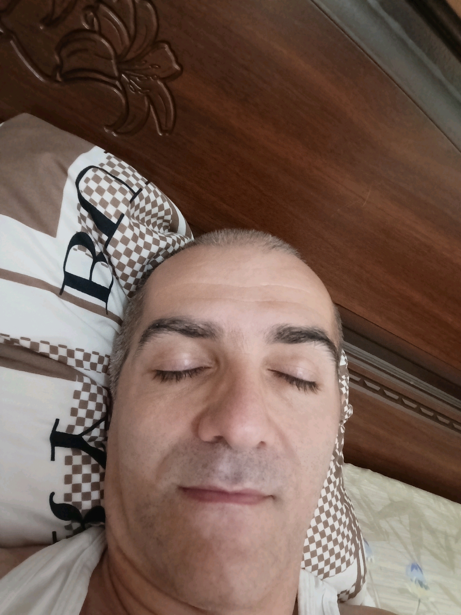 An overhead shot captures a man lying in bed with his eyes closed, facing upwards towards a wooden headboard. He has short, grey hair and appears to be in his 40s or 50s. He is wearing a white shirt. The bed is covered with a pillow featuring a checkered brown and white pattern and floral motifs. The headboard is made of dark wood and has an ornate carved design of a flower. The room appears to be a bedroom, likely in a residential setting in Grozny, Russia. The lighting suggests it is daytime.