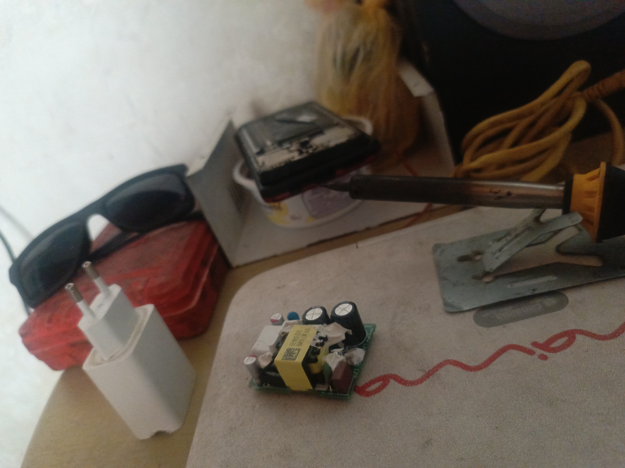 The image shows a cluttered workbench or table. Prominently featured is an electronic circuit board, likely from a power adapter, with visible components like capacitors and a yellow transformer. Next to it is a white power adapter. A soldering iron is present, along with a soldering stand. Other objects in the scene include a pair of sunglasses, a red toolbox, a container with a dark object inside, and a yellow cord. The work surface has a pattern in red ink.
There is no information to determine if the location is in Juan Griego, Venezuela.