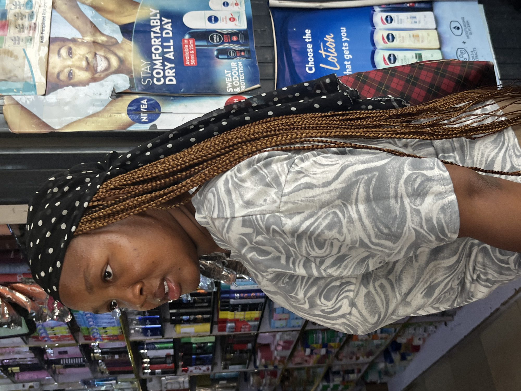 The image shows a young Black woman from the side, wearing a black headscarf with white polka dots and a grey and white patterned shirt. She has long braided hair. She is standing in front of a store display. The store appears to be a pharmacy or cosmetic shop, with shelves filled with various products. In the background, there are posters advertising Nivea products, including deodorant and lotion. The poster for lotion says "Choose the Lotion that gets you". The woman is looking to her left, with her mouth slightly open. The environment suggests an urban setting, likely a commercial area within the city of Jos, Nigeria, given the woman's appearance and the typical layout of shops in that region.