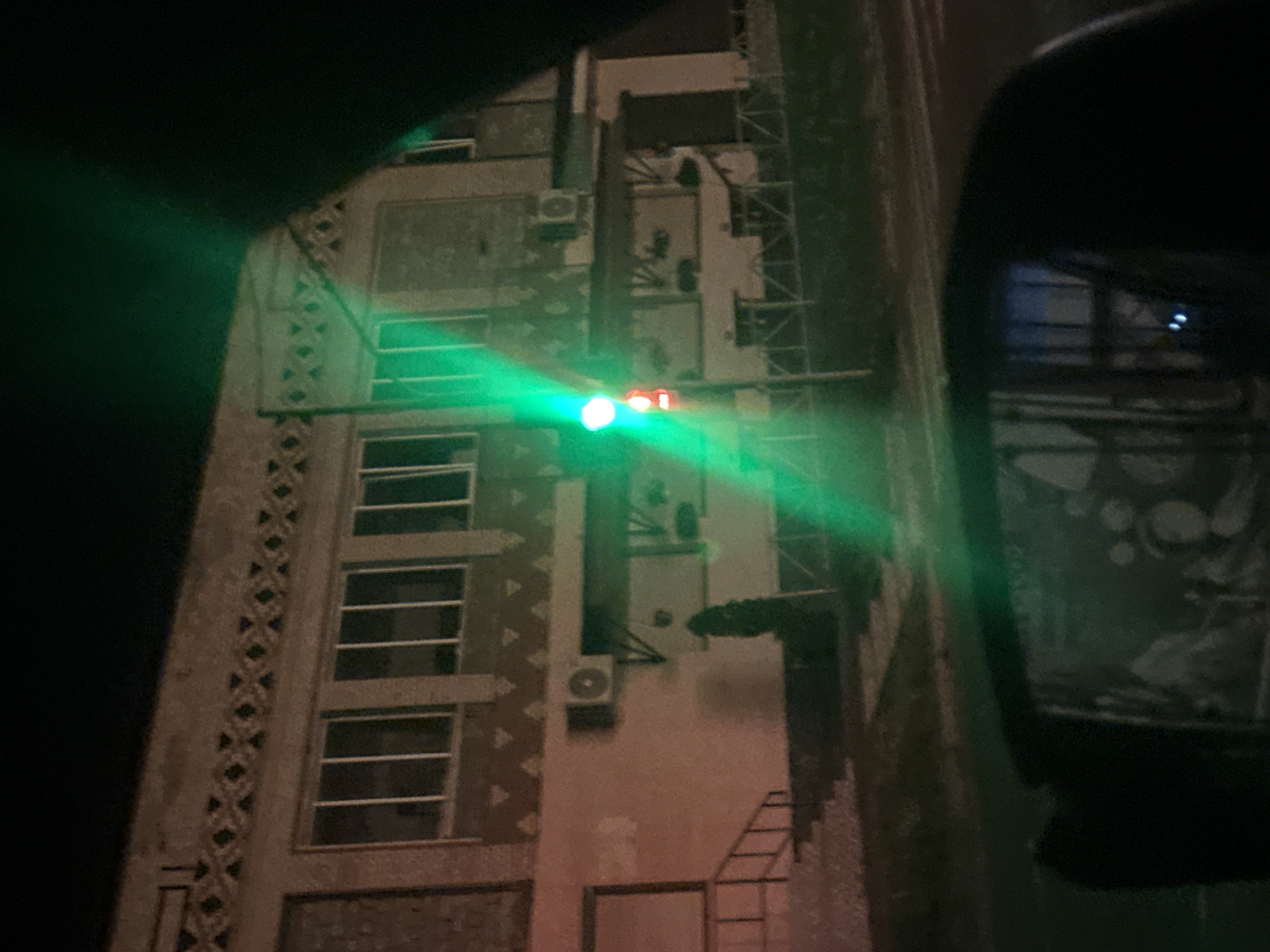In Shatava, Ukraine, at night, a green traffic light illuminates a building with a textured facade and barred windows. The building appears to be a commercial or industrial structure, possibly abandoned, with an awning covering a storefront. A black car's side mirror is visible in the foreground, reflecting the building's interior and a portion of the street. The overall scene is dark and somewhat eerie, with the green light casting long shadows and creating a sense of mystery.