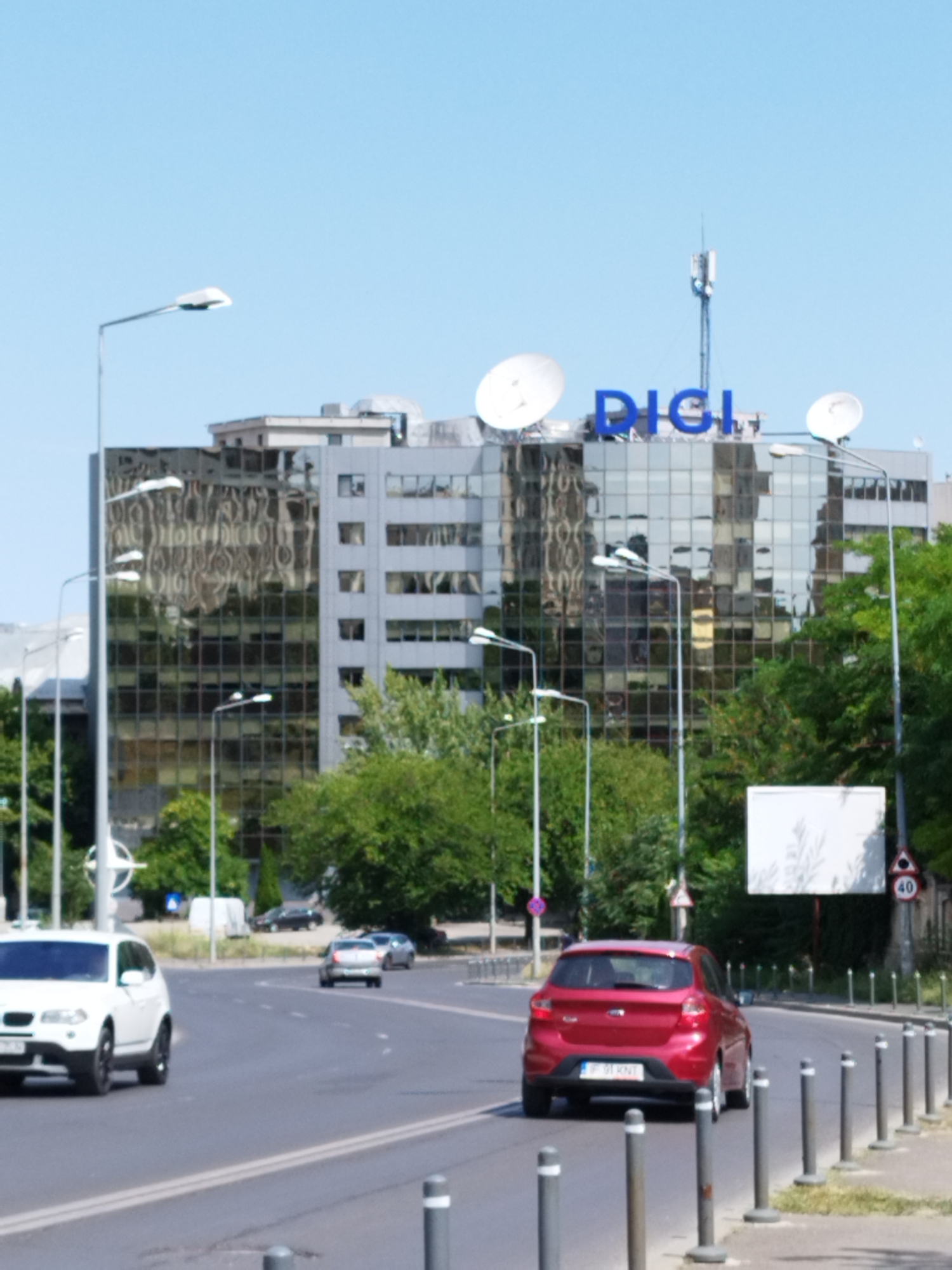 The image shows a street scene in Bucharest, Romania, with a prominent glass-facade building bearing the blue "DIGI" logo and satellite dishes on its roof. A white SUV is parked on the left side of the road, while a red hatchback is driving away in the right lane. The street is lined with trees and lampposts, and there are other cars and a billboard visible in the background. The sky is clear and blue.