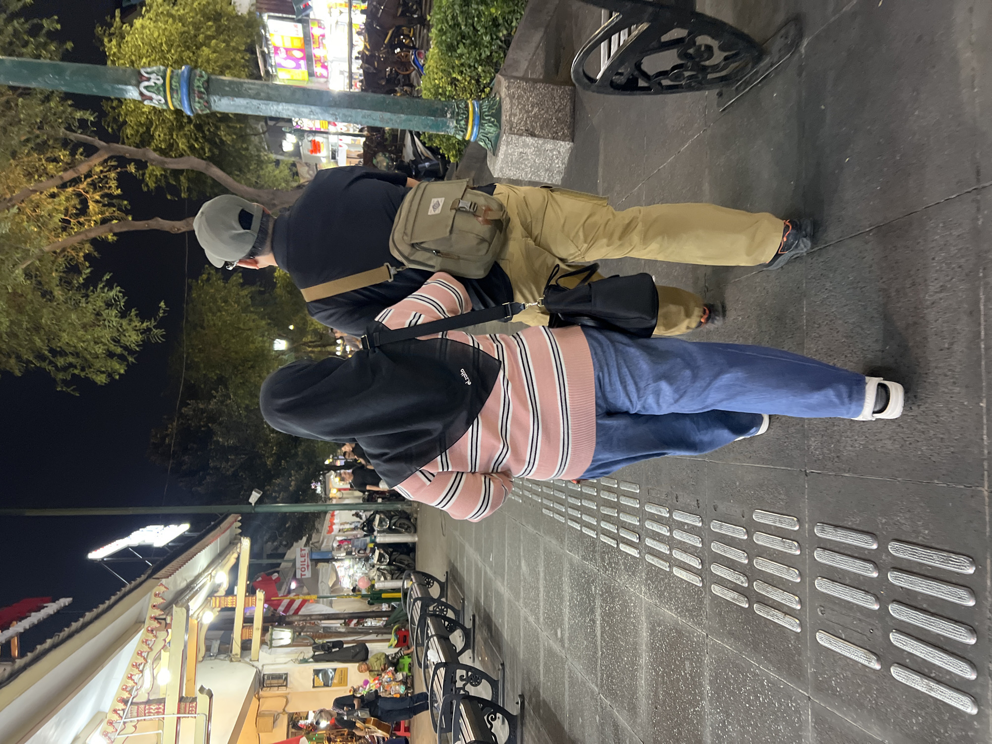 Two individuals, a woman wearing a black hijab and a pink and white striped sweater with blue jeans, and a man wearing a black shirt, tan cargo pants, and a grey baseball cap, walk away from the camera on a cobblestone sidewalk at night. The woman carries a black handbag. The man has a tan crossbody bag. They are walking on a street in Yogyakarta, Indonesia, indicated by a sign for "TOILET" and storefronts with colorful awnings and lights. Several benches are visible along the sidewalk. The ground is marked with tactile paving for the visually impaired. The atmosphere appears to be a lively urban commercial area at night.