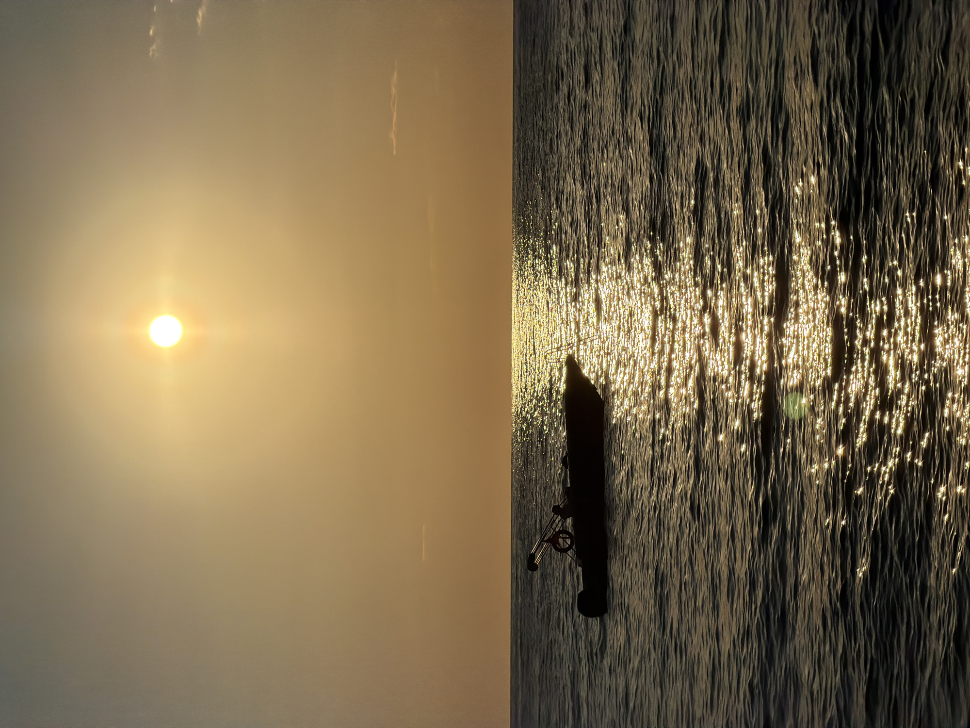 The image shows a scene at sunset, likely in Fethiye, Turkey. The left side displays the sun in a hazy sky. The right side depicts water with the sun reflecting off the surface, creating a shimmering effect. A small boat with a fishing rod is silhouetted against the water. The image is rotated 90 degrees counter-clockwise.