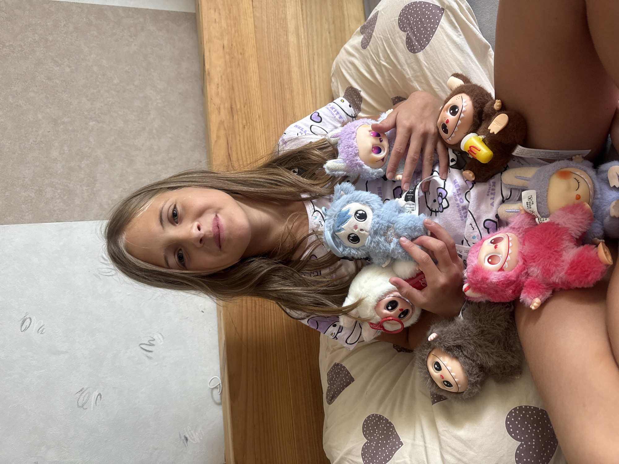 A young girl with long brown hair lies on a bed, surrounded by several plush toys. She is wearing pajamas with a heart pattern. The toys include a brown monkey with a yellow cup, a light blue creature, a fluffy white creature with large eyes, a gray furry creature, and a pink creature. The girl is looking at the camera with a slight smile. The bed appears to be made of wood, and the room is lit, suggesting daytime. The girl's relaxed pose and the presence of numerous toys indicate a casual, possibly playful moment. The background includes a wall with a textured pattern and what seems to be a wooden headboard. Some of the toys have visible tags.