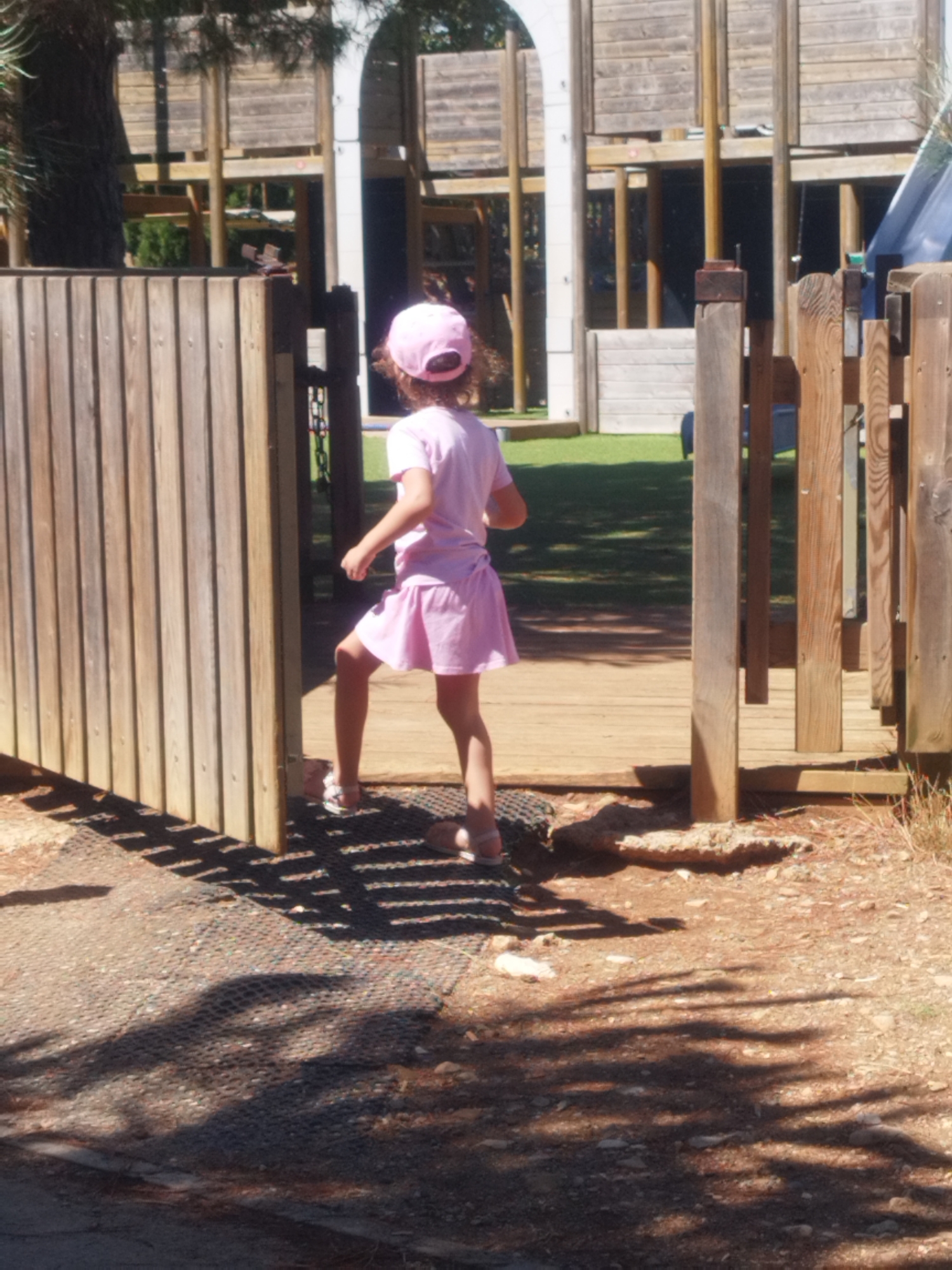 In Antibes, France, a young girl wearing a pink cap and a pink dress is captured mid-stride, ascending a wooden ramp within a playground. Her right leg is elevated as she moves forward. The playground features rustic wooden structures, including a fence to her left and a series of vertical wooden posts to her right, forming an entryway. Sunlight casts a dappled shadow pattern on the ground, a mix of gravel and a dark, mesh-like surface. The background reveals more of the playground's construction, with tiered wooden platforms and supports, and hints of greenery beyond.