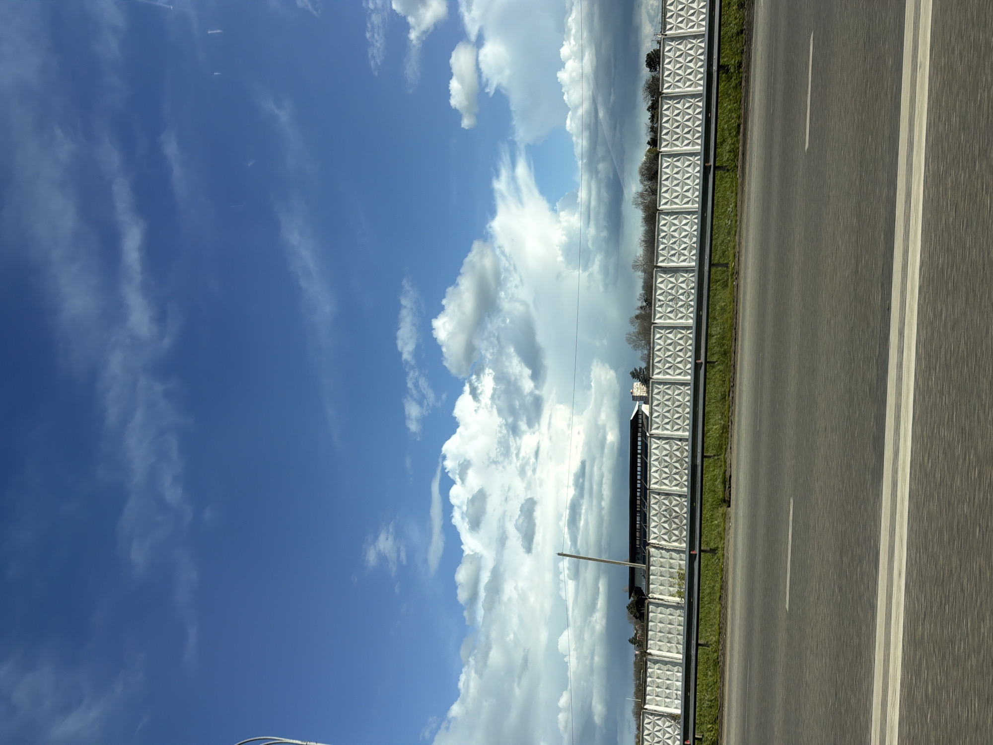 The image depicts a highway in Lazurny, Russia. To the right is an asphalt road with two lanes separated by a solid white line. A concrete barrier runs parallel to the road, decorated with a pattern of geometric shapes. Beyond the barrier, there is greenery and some trees. To the left, a vibrant blue sky filled with white clouds dominates the scene. A utility pole and wires are visible amidst the clouds.